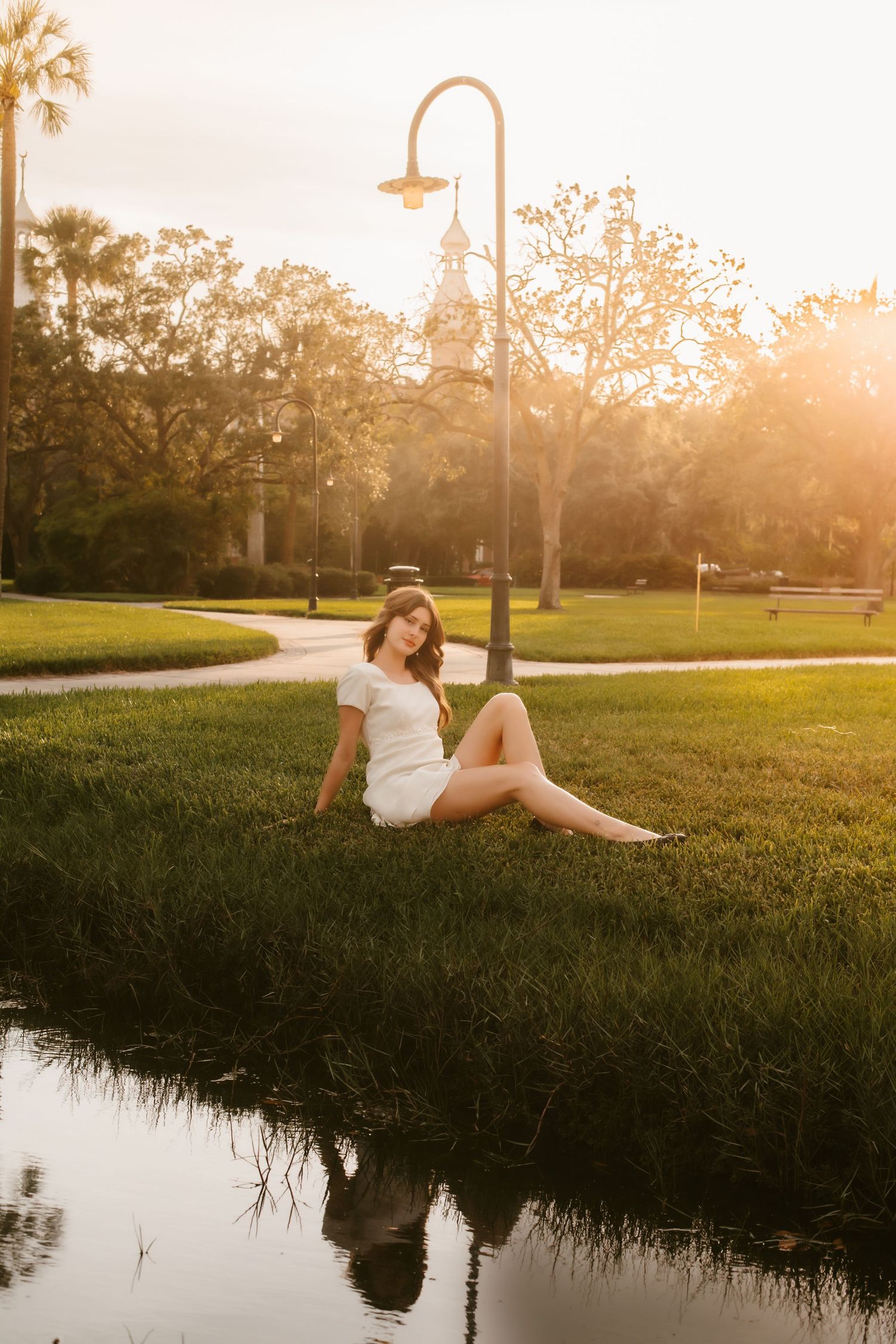 Young woman in white dress sitting on grass by a pond at golden hour sunset in a lush green park.