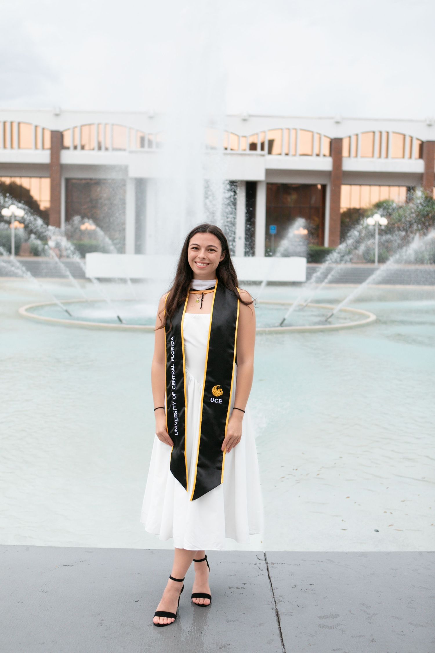 Smiling female graduate in white dress and UCF black stole poses by fountain on campus.