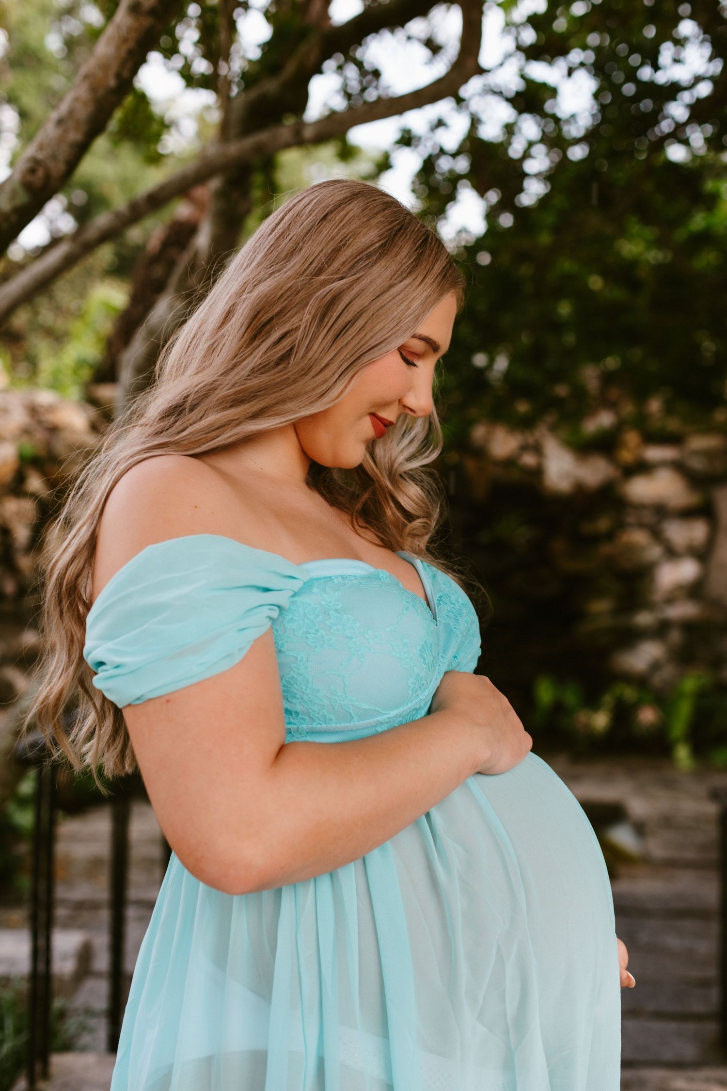 Pregnant woman in a teal off-shoulder gown cradling her baby bump outdoors among lush greenery.