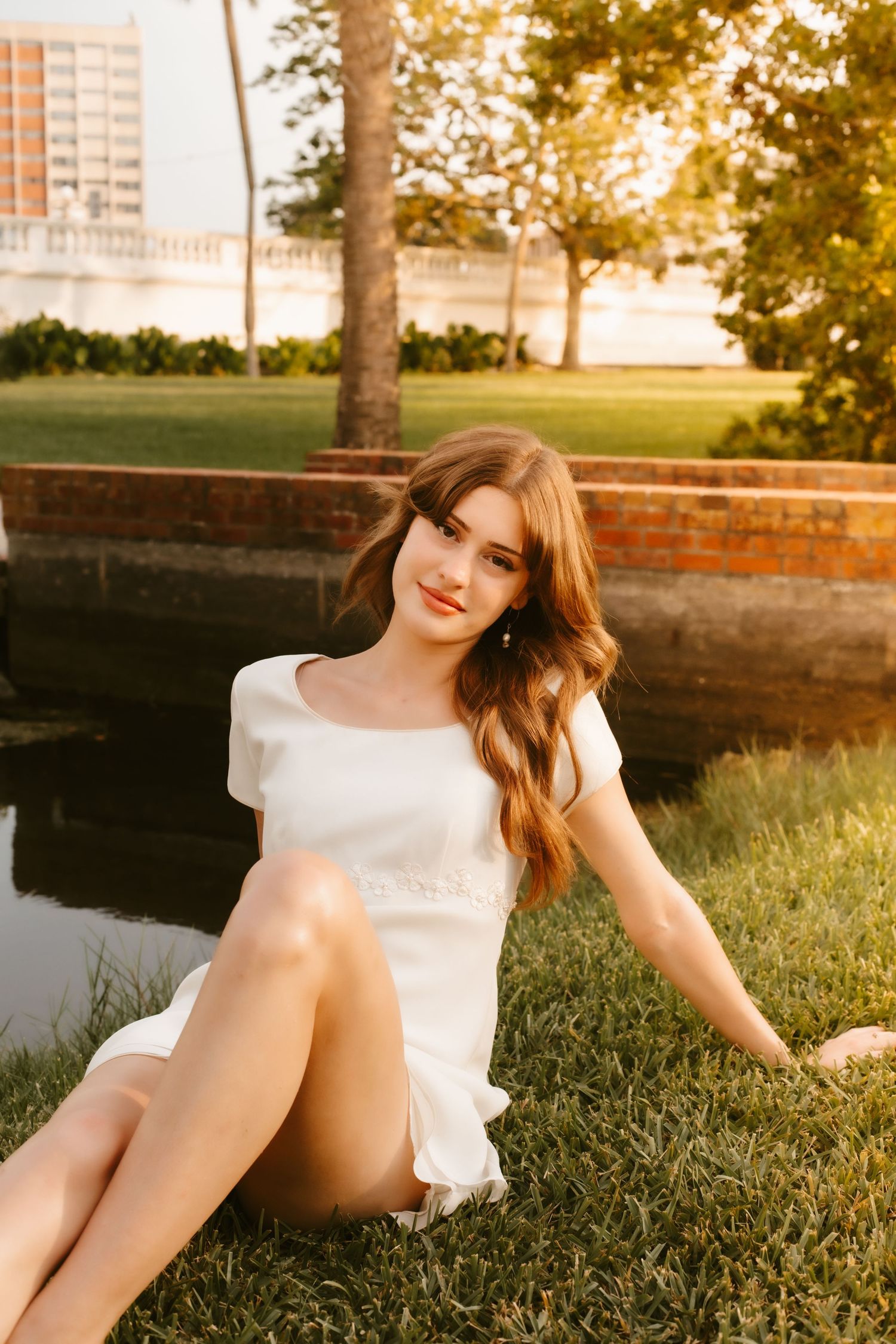 Smiling young woman in white mini dress sitting on grass by a pond in a warm golden hour park setting.