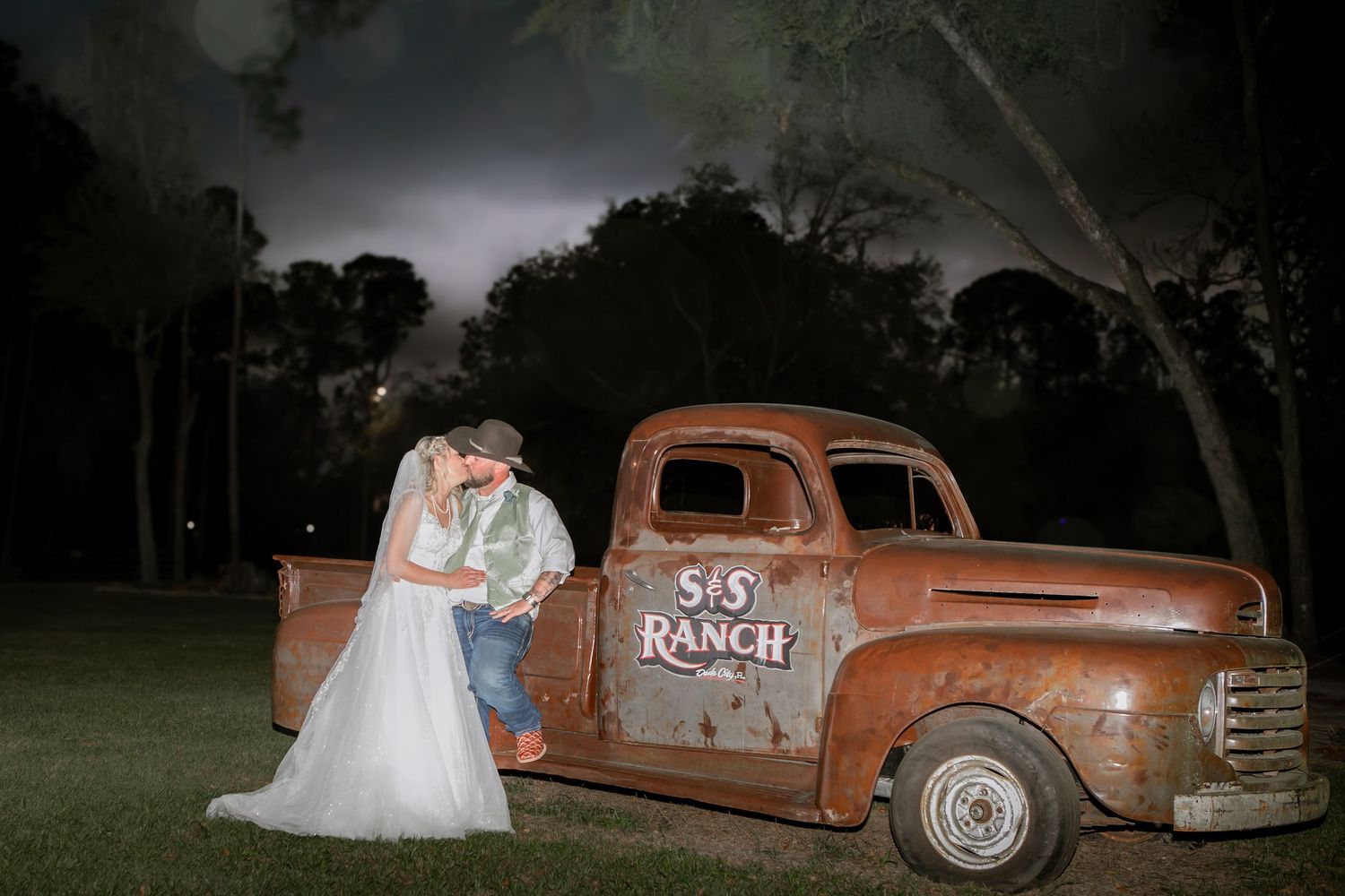 Bride and groom share a romantic kiss beside a vintage rusty S&S Ranch pickup truck at night.