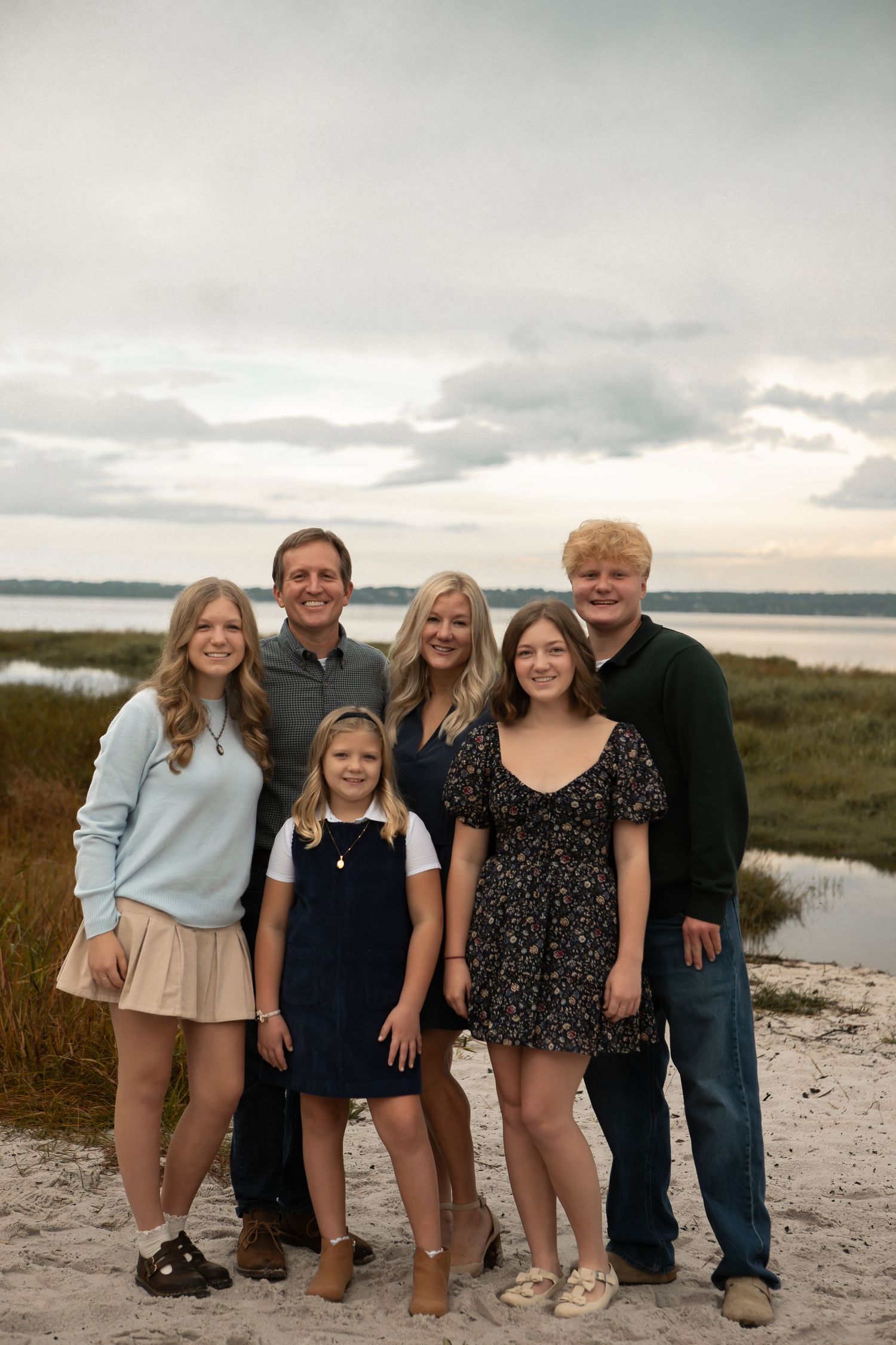 A family of six poses together on a sandy beach with a scenic marsh and cloudy sky.