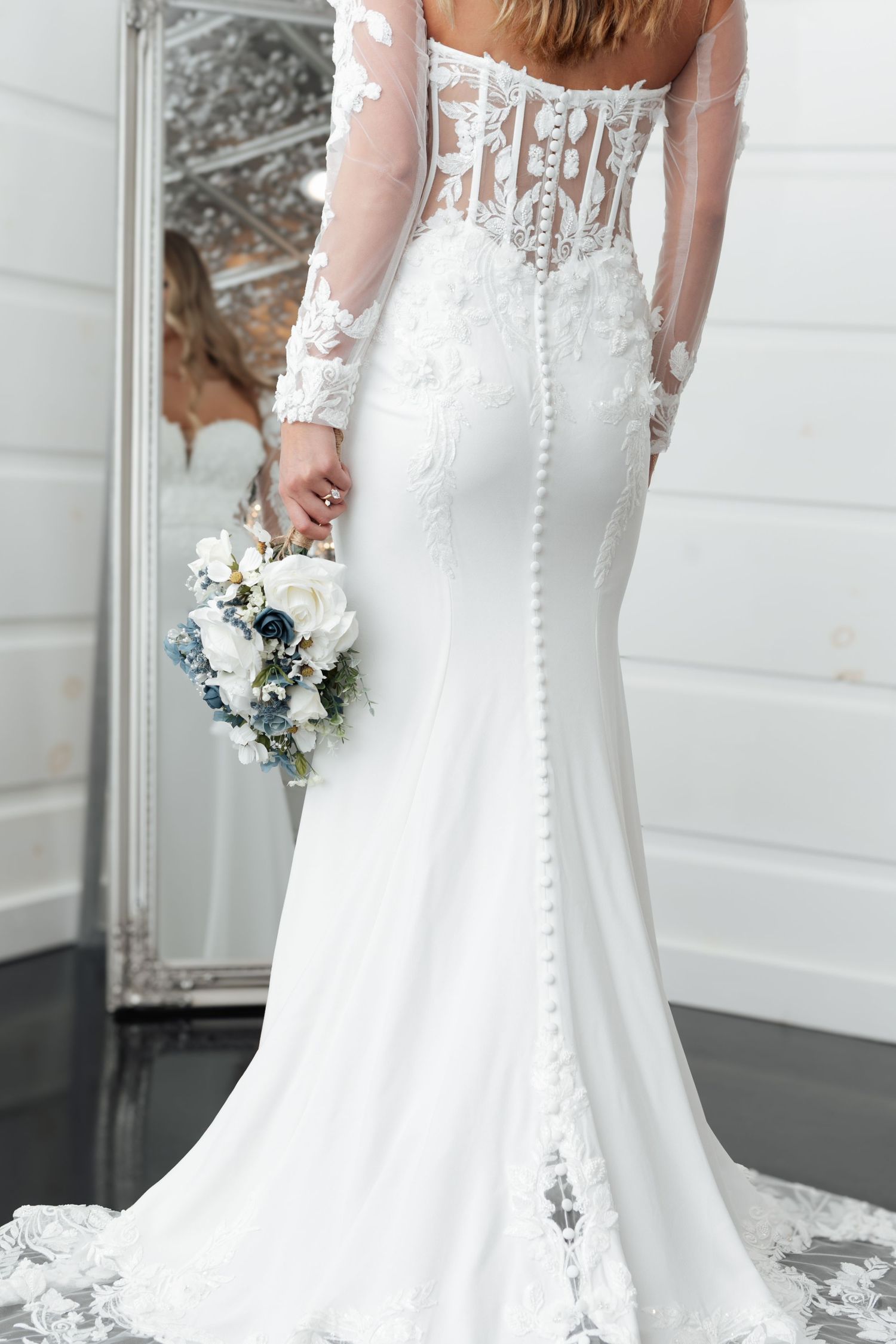 Bride in elegant white lace mermaid wedding gown with sheer back and long sleeves holding a blue and white bouquet.