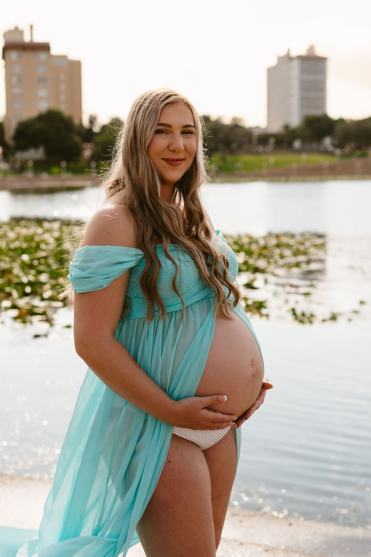 Pregnant woman in teal sheer dress holding baby bump near a serene lake with city buildings at sunset.