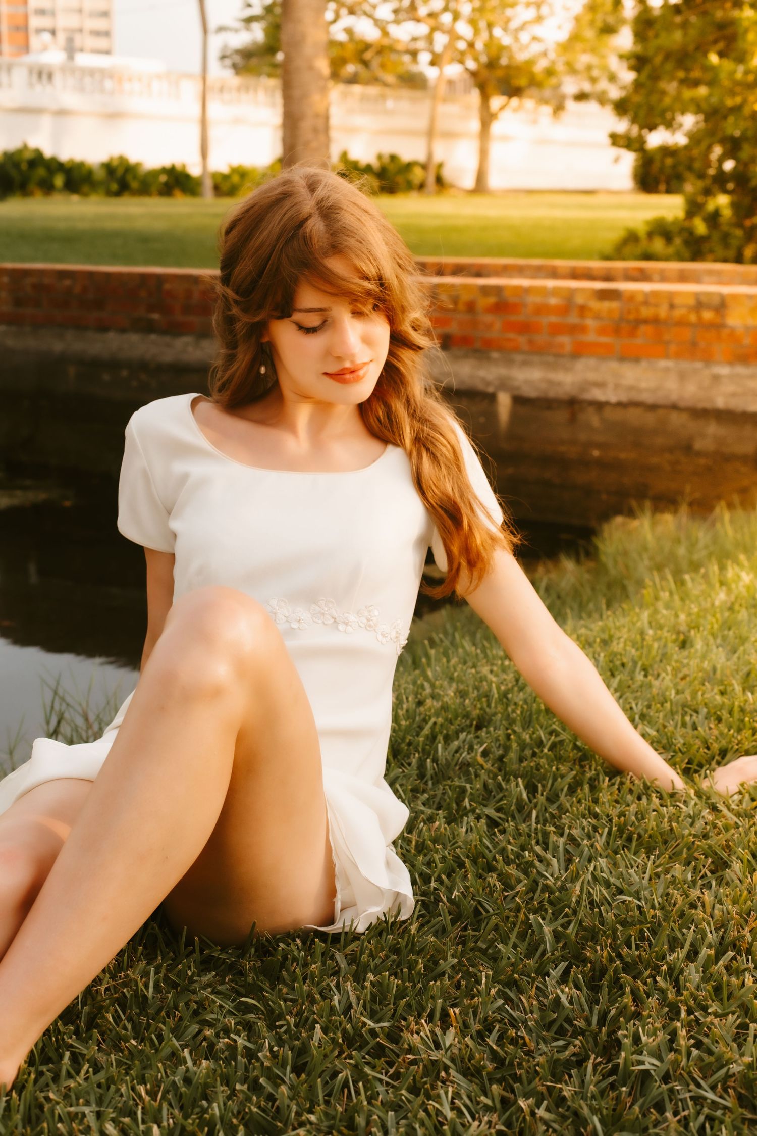 Young woman in white mini dress sitting on grass by a pond at golden hour in a park setting.