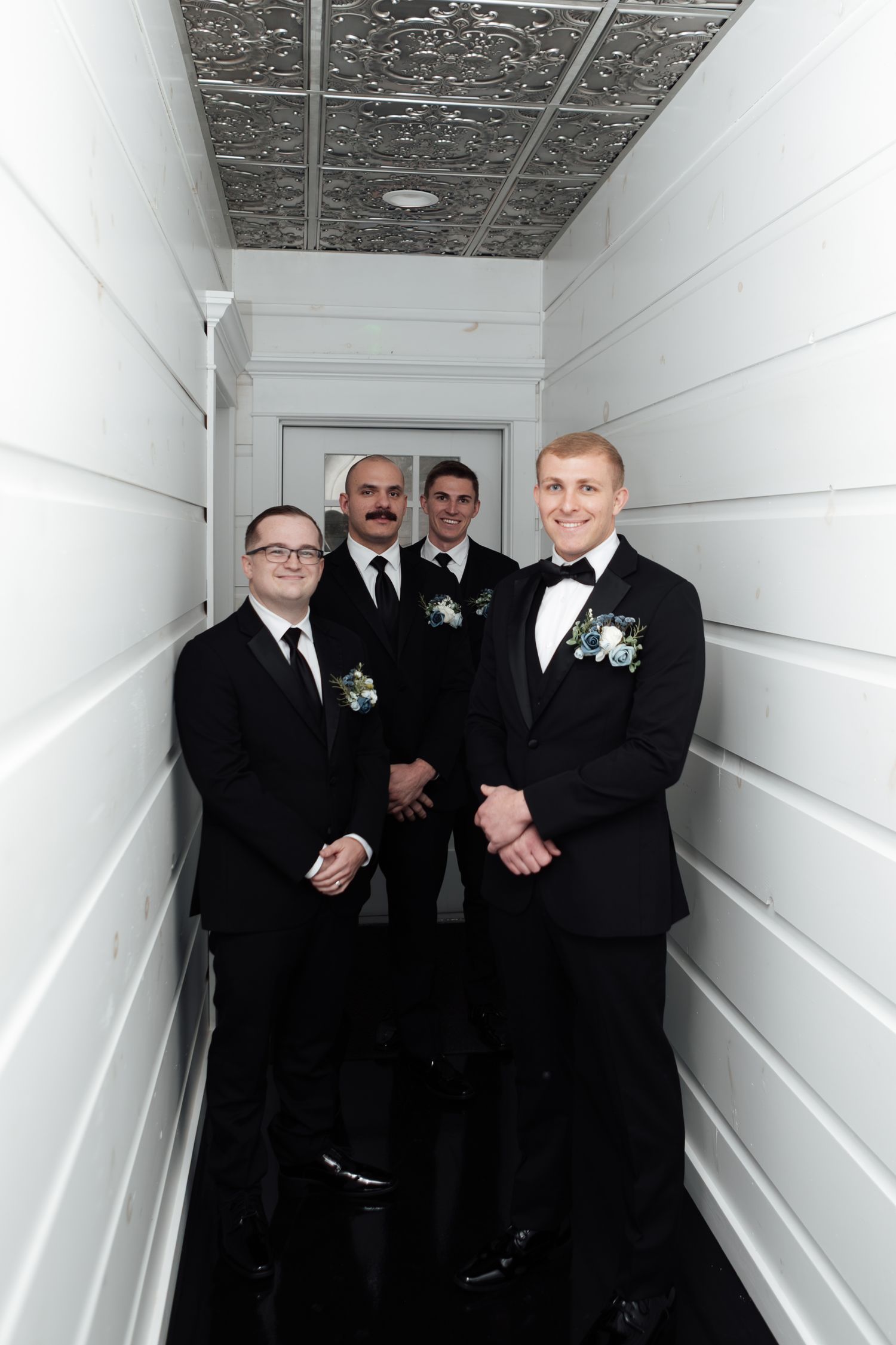 Four groomsmen in black tuxedos with boutonnieres posing in a white hallway before a wedding ceremony.