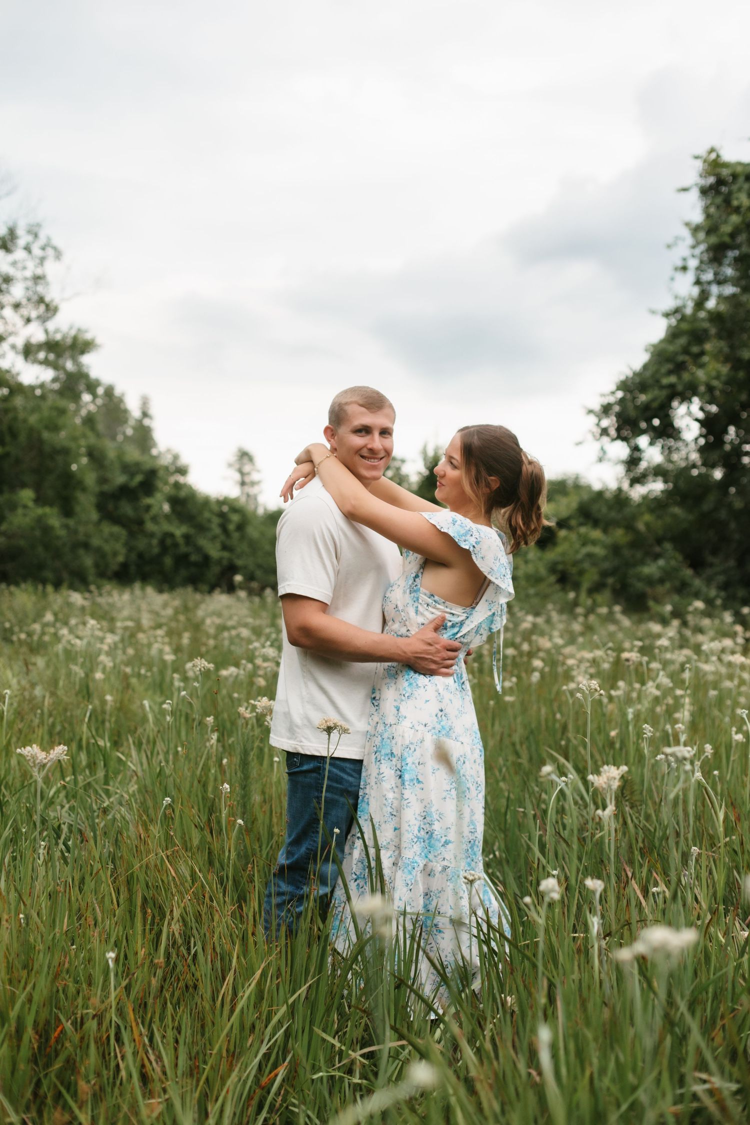 Happy couple embracing and smiling in a field of white wildflowers, woman in blue floral dress with arms around mans neck.