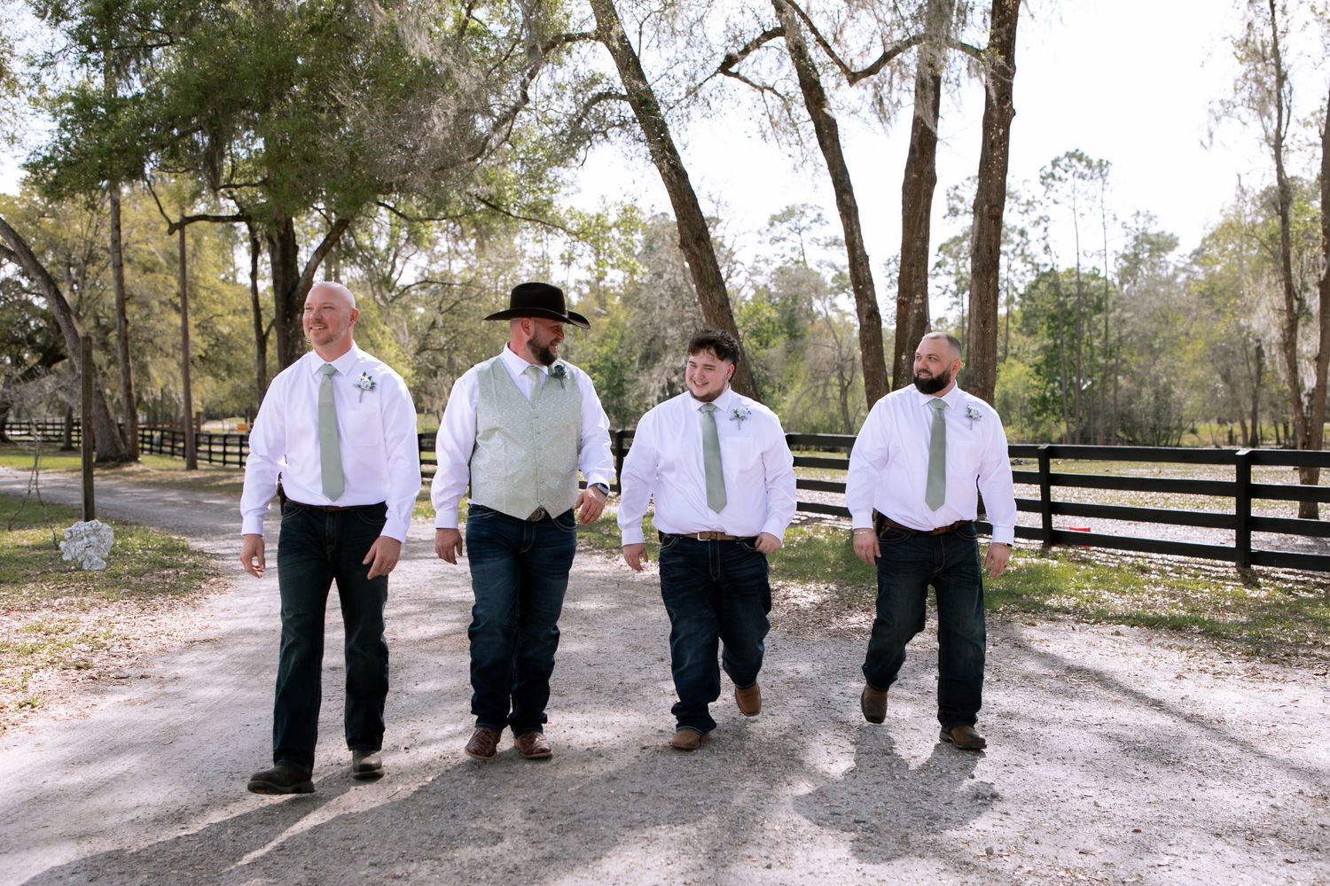 Groom and groomsmen walking together in casual western attire along a tree-lined ranch driveway.