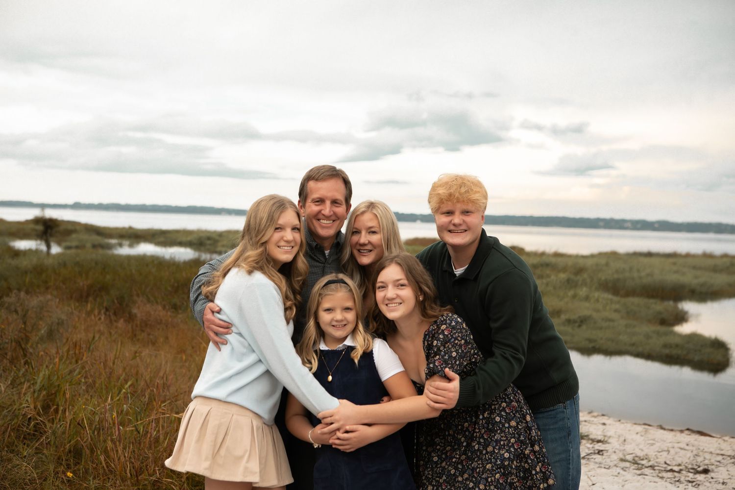 Happy family of five poses together outdoors near a scenic waterway under cloudy skies.