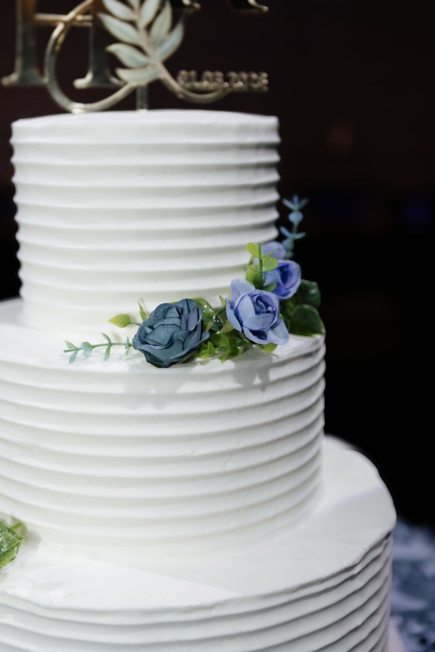 Elegant three-tier white ribbed wedding cake decorated with blue and green floral accents.