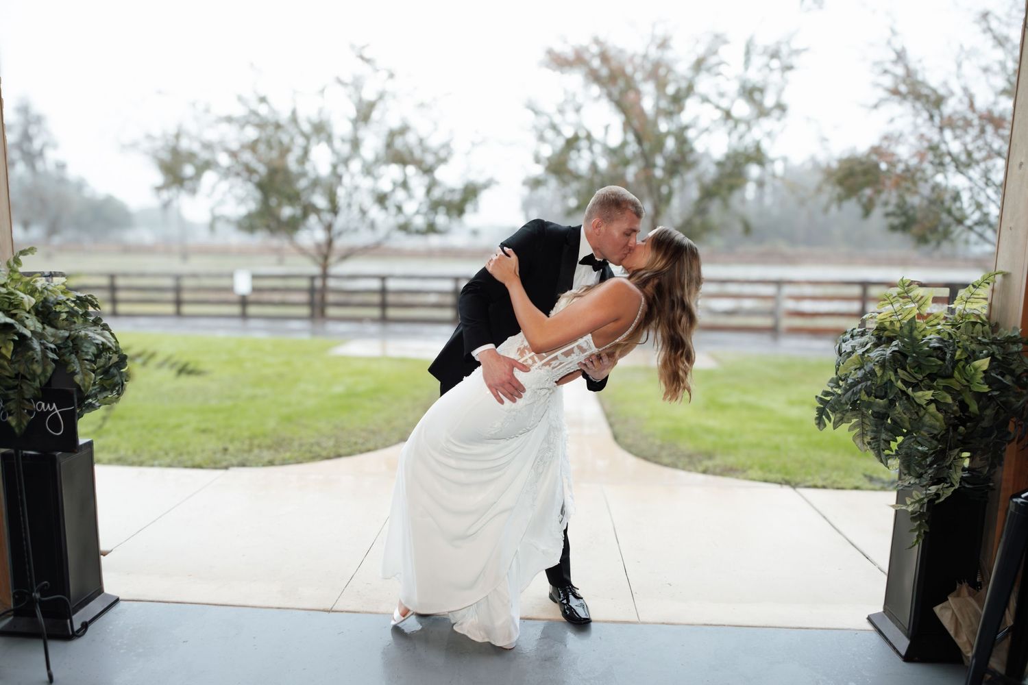 Groom dipping bride for a romantic kiss outdoors at a rustic wedding venue with lush greenery.