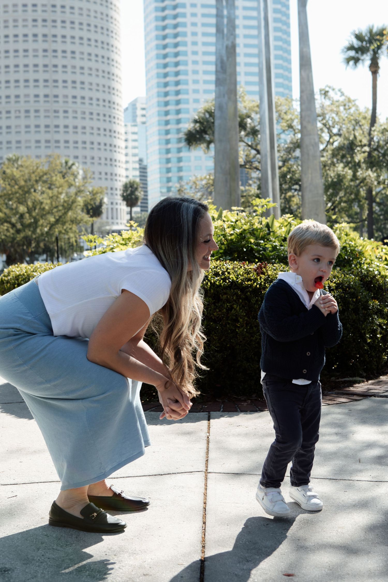 Woman bends down to interact with a toddler eating a lollipop in an urban park setting.