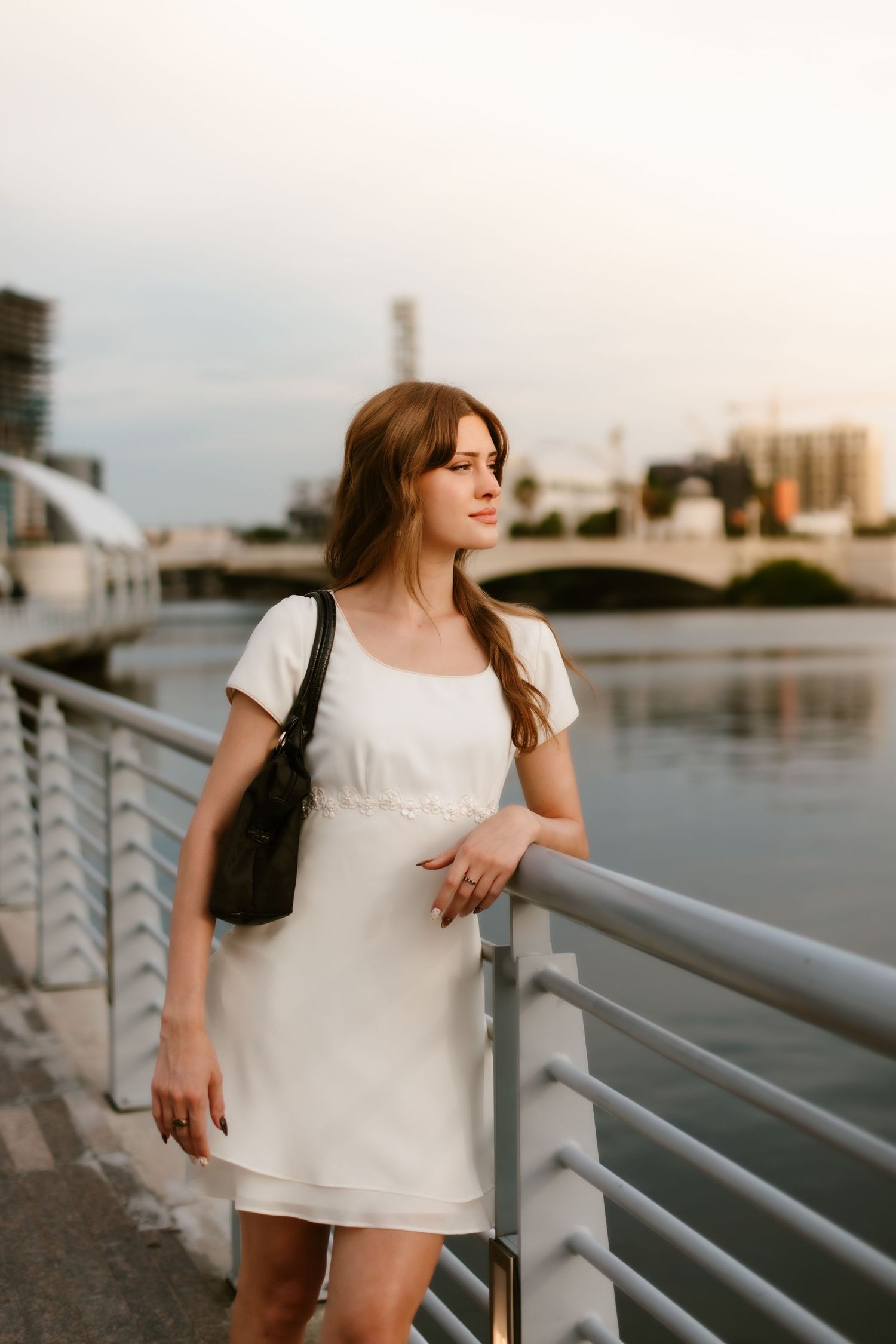 Young woman in white dress with black bag leaning on railing by urban waterfront at sunset.