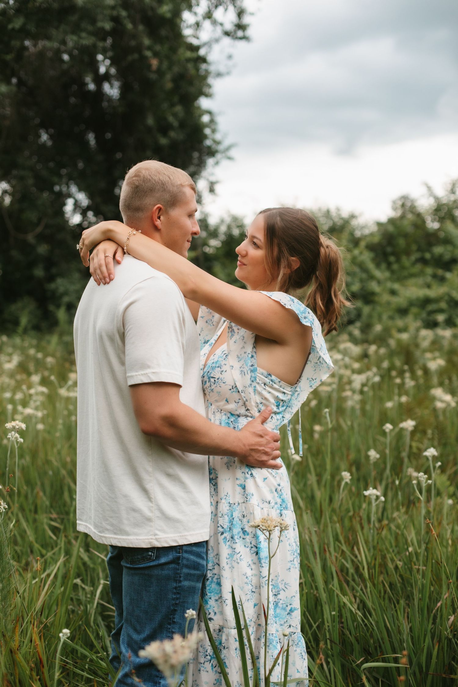 Couple gazing into each others eyes while embracing in a green meadow filled with white wildflowers.