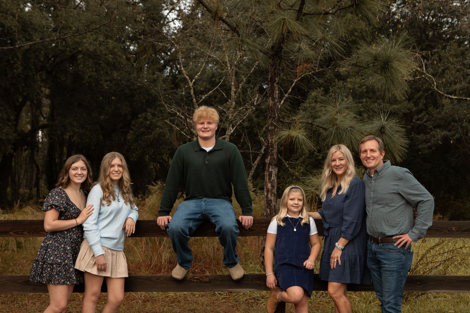 A family of six poses outdoors in a wooded setting with a young man seated on a fence.