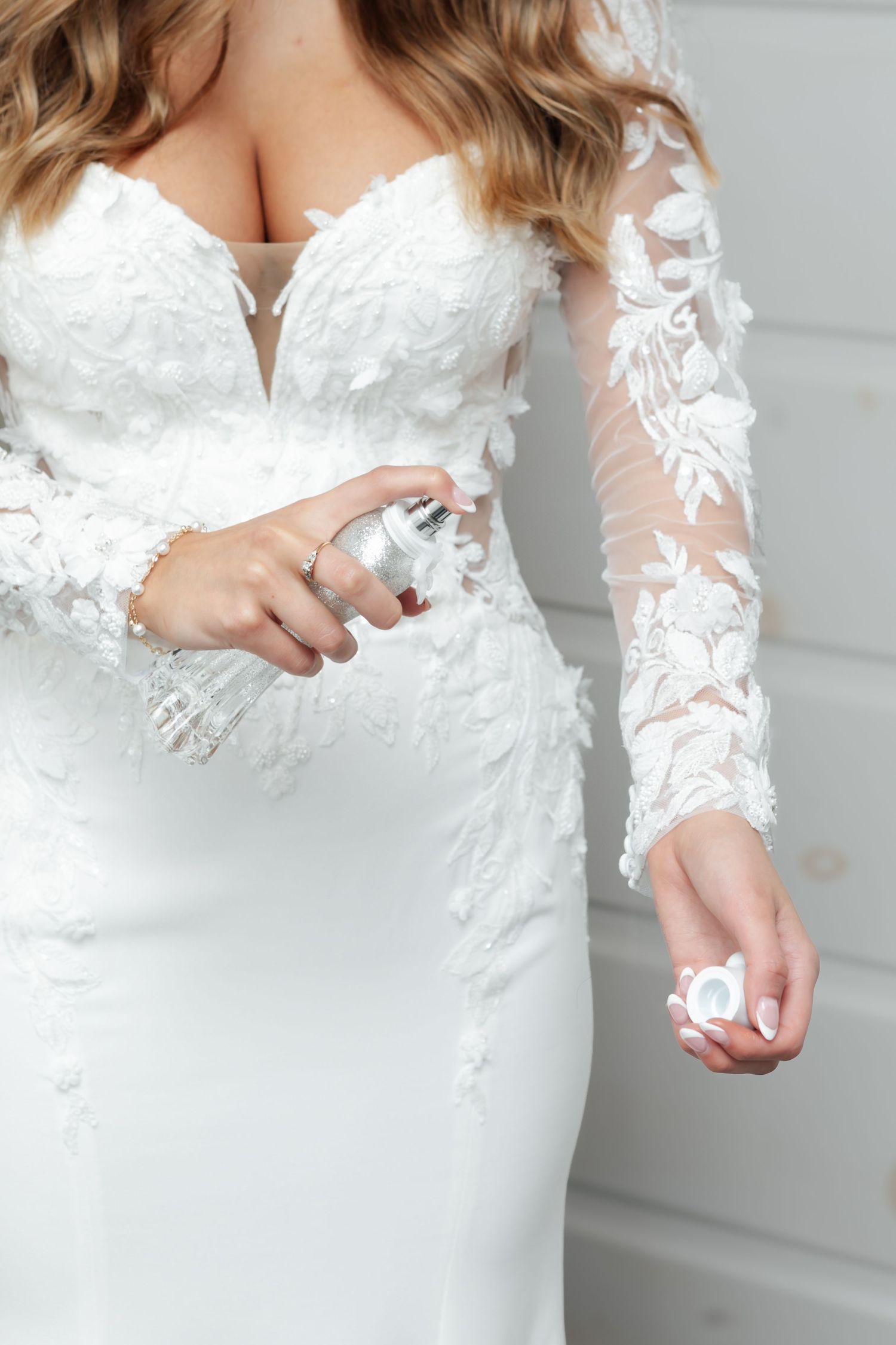 Close-up of bride in white lace long-sleeve wedding gown holding a perfume bottle with elegant floral applique detail.