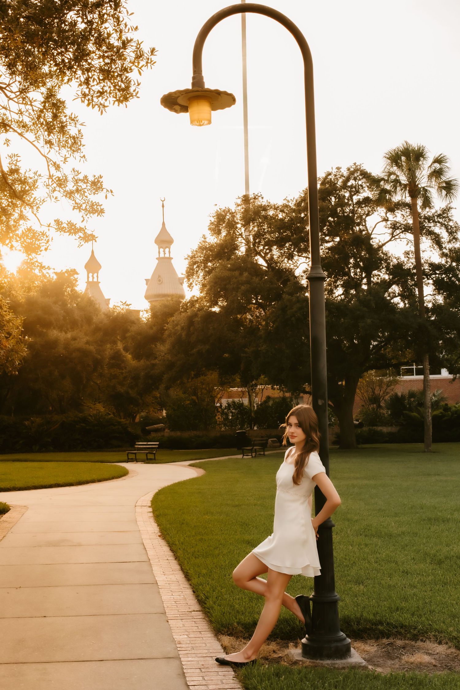 Young woman in white dress leans against lamp post on brick path at sunset near historic building with minarets.