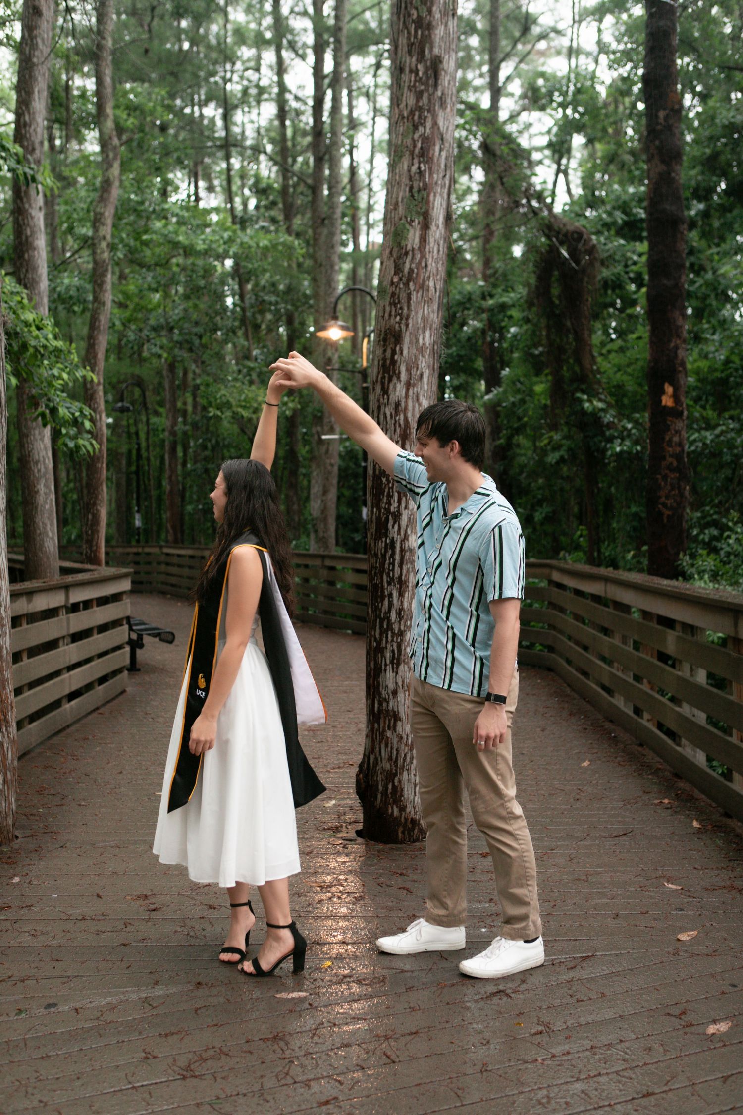 Couple dancing on a wooden boardwalk in a pine forest with string lights overhead.
