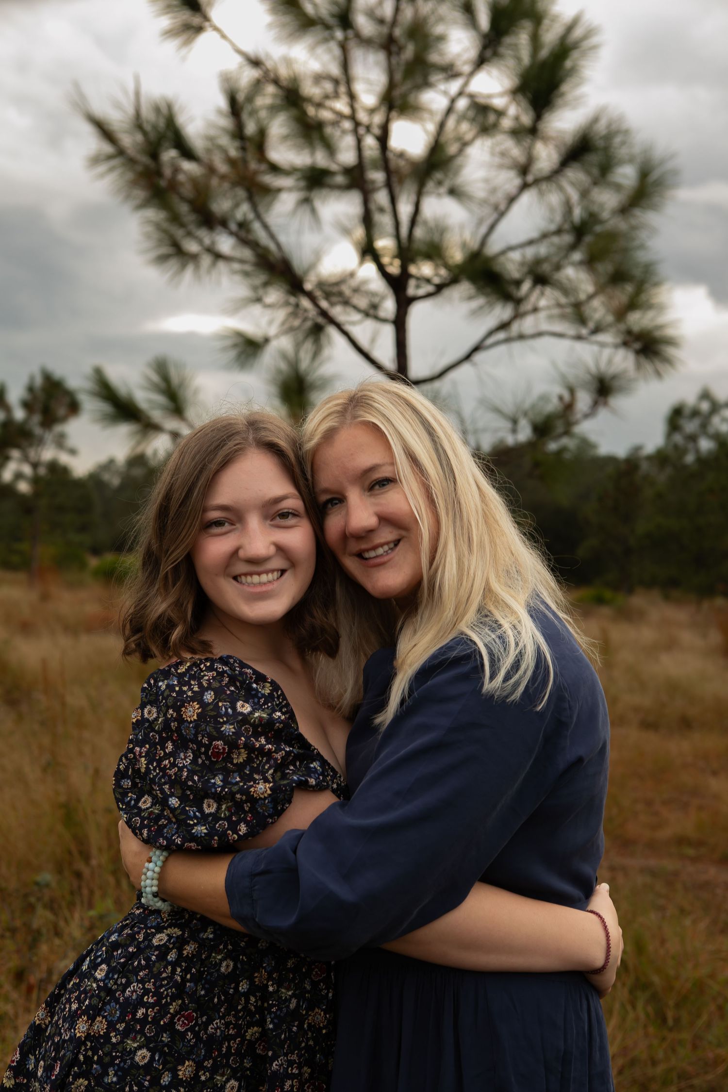 Two smiling women embrace warmly outdoors in a field with a pine tree in the background.