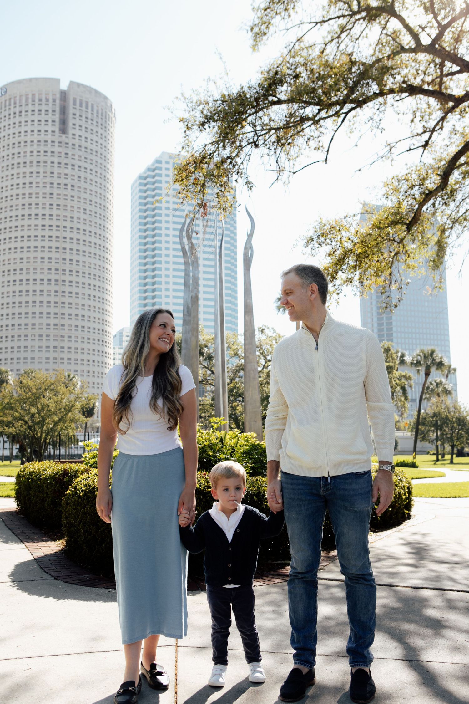 Young family of three holds hands smiling in Tampa park with downtown skyscrapers in background.