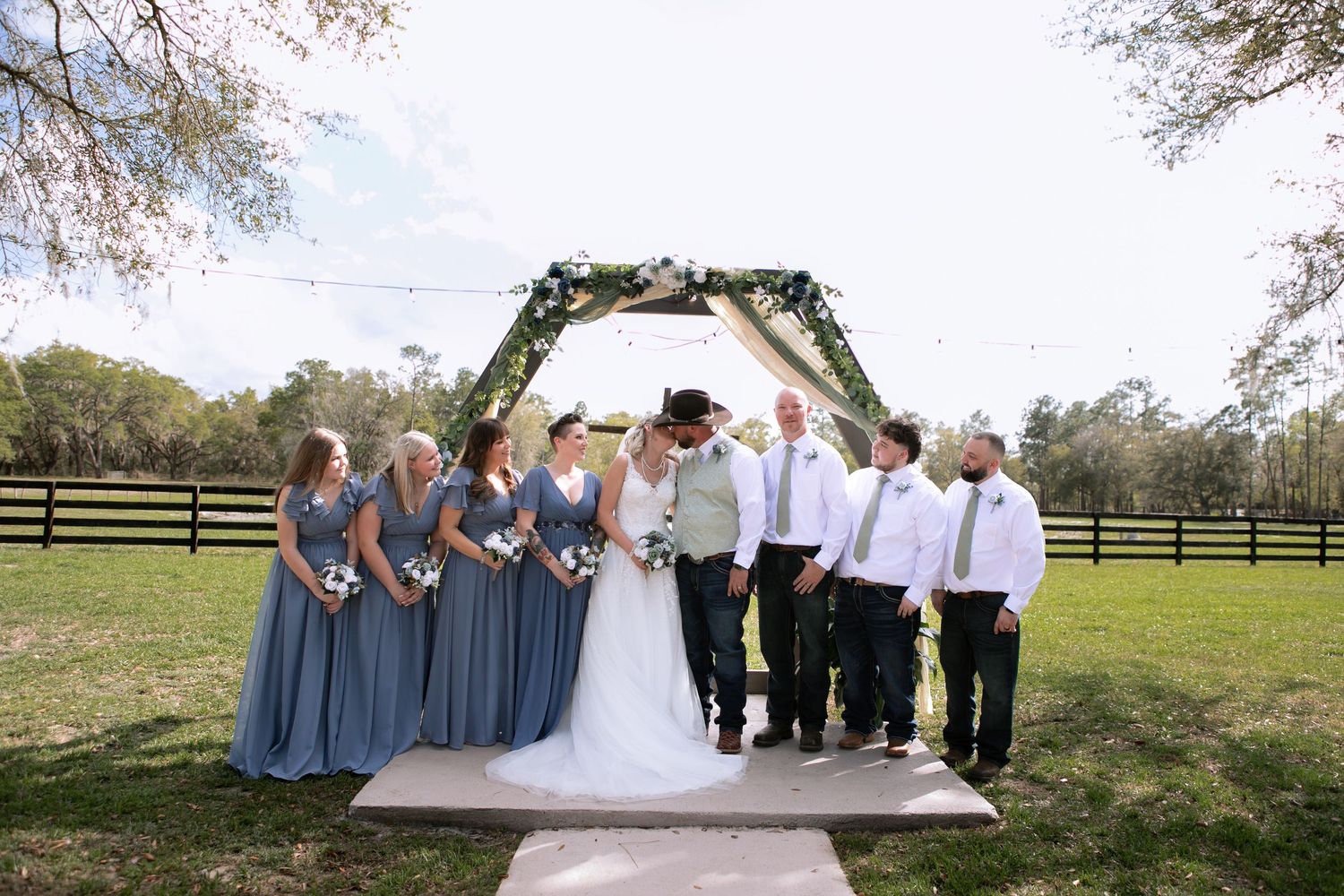 Wedding party poses under a hexagonal floral arch on a green lawn, bridesmaids in blue dresses with groomsmen.