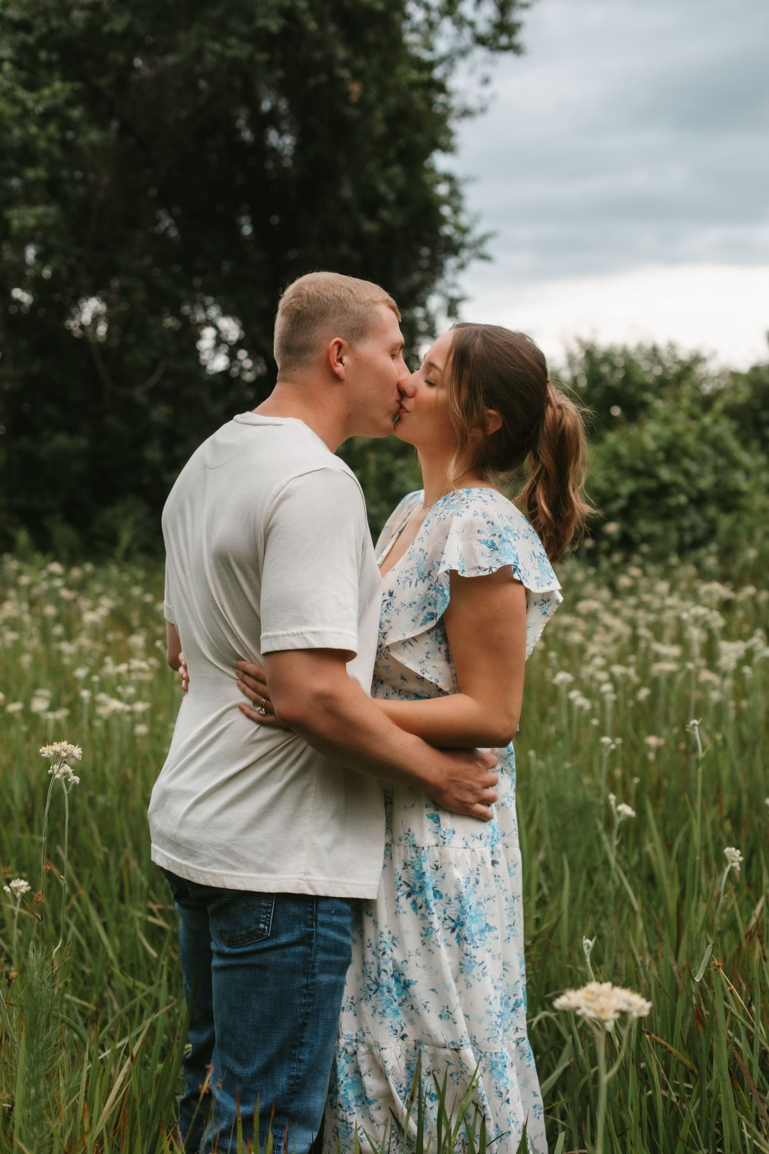 Couple sharing a kiss in a wildflower field, woman in blue floral dress embraced by man in white tee.