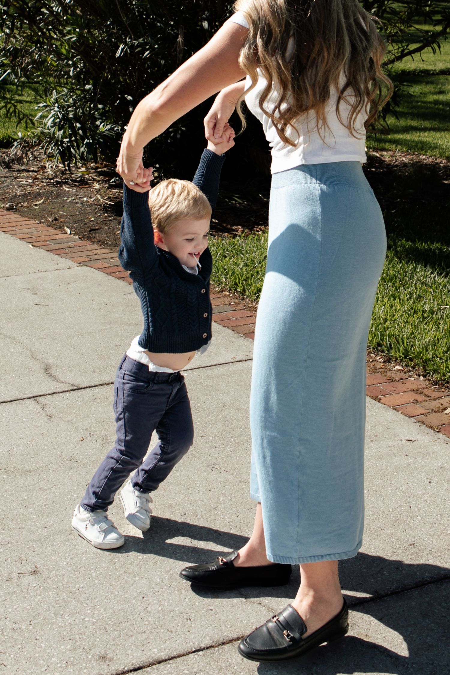 Joyful young boy laughs as his mother playfully spins him by his hands on a sunny outdoor sidewalk.