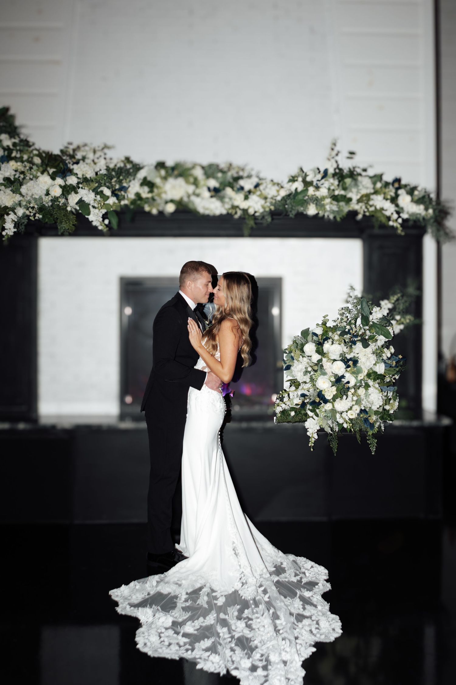 Bride and groom sharing a romantic embrace in front of a white floral fireplace display.