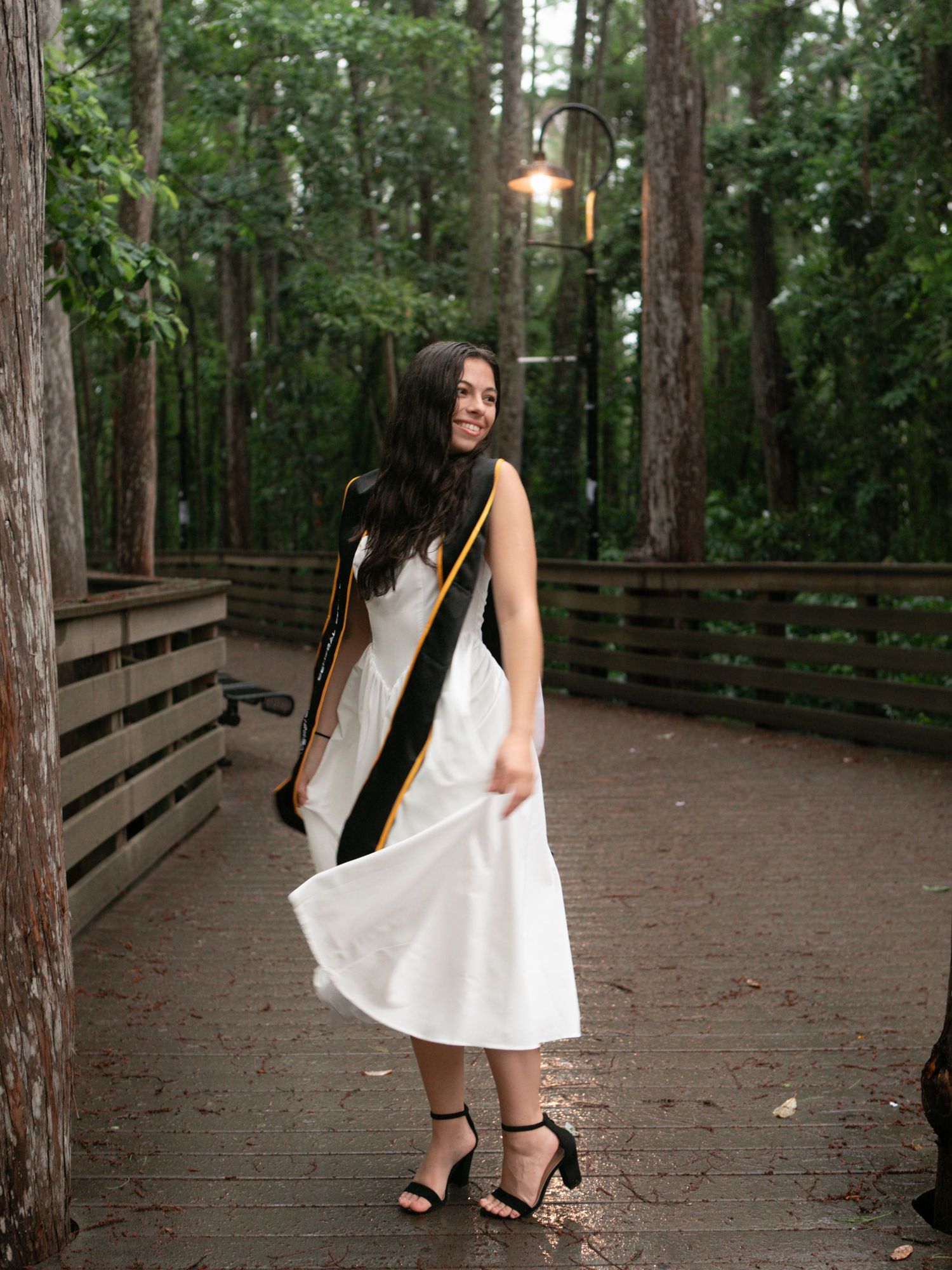 Female graduate in white midi dress and black stole twirls on wooded deck path smiling.