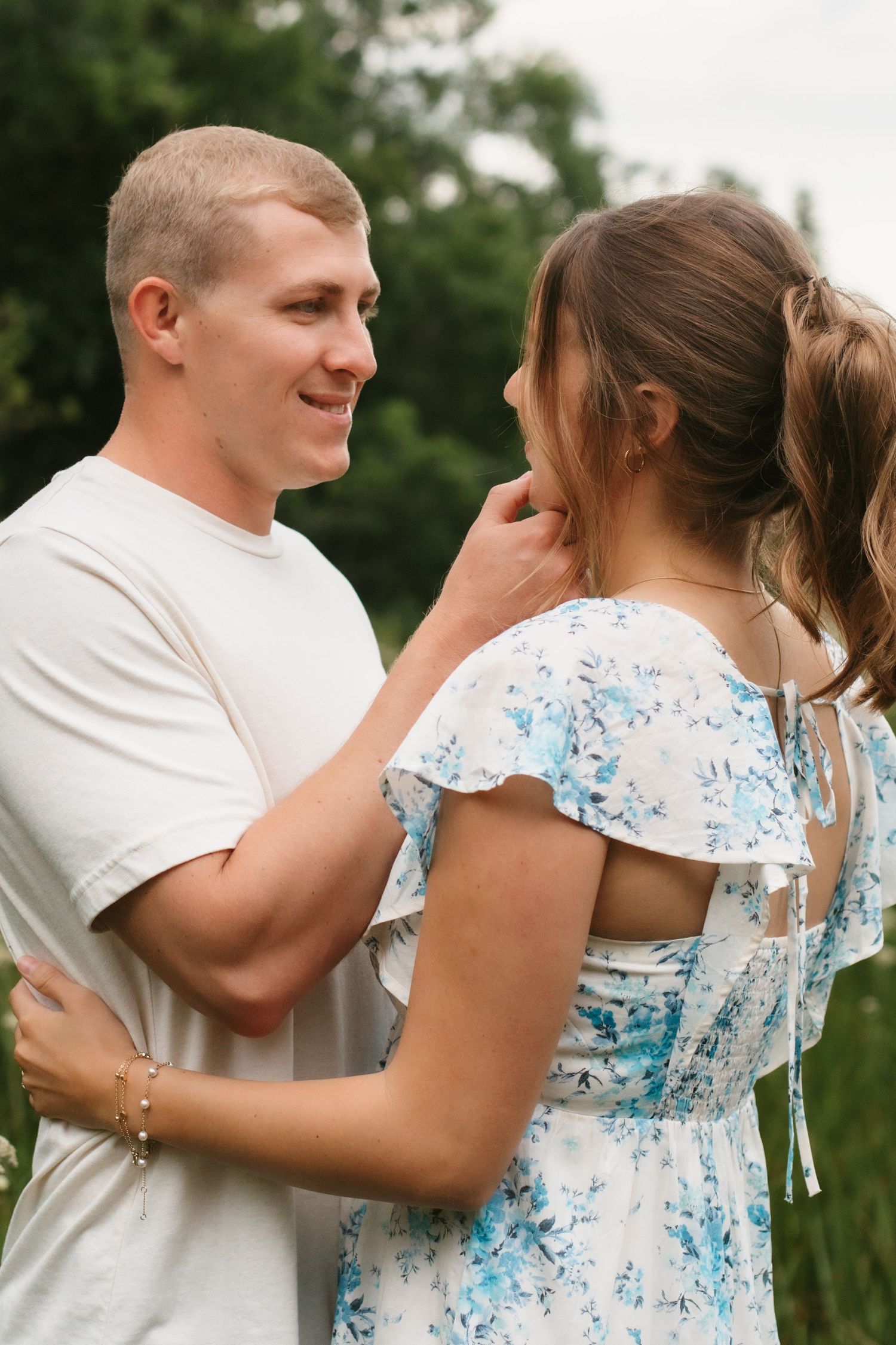 A smiling couple embraces outdoors, man in white tee touches woman's hair as she wears a floral blue dress.
