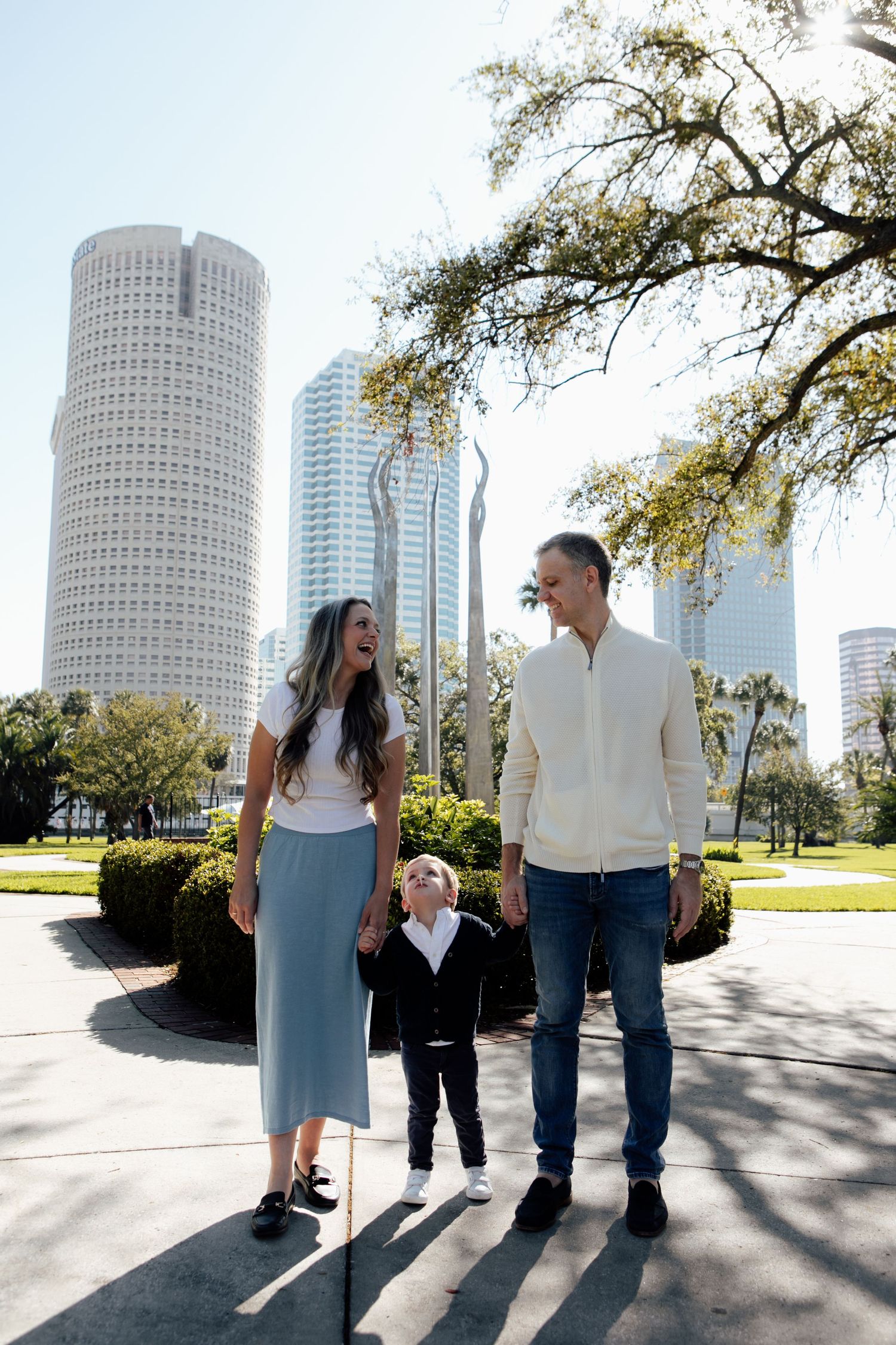 Happy young family with toddler son walks together in a sunny urban park with city skyscrapers in the background.