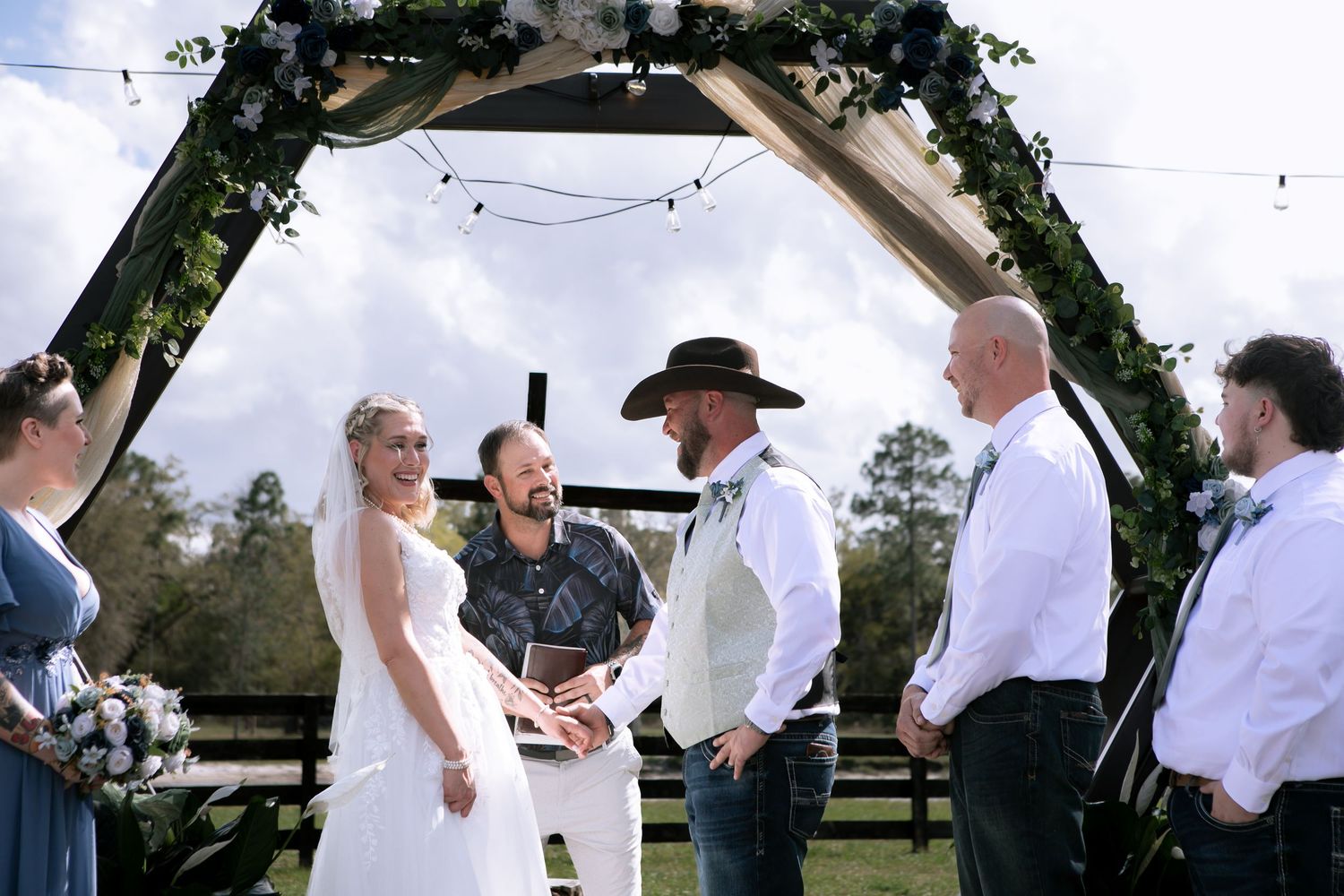Bride and groom exchange vows under a floral hexagonal arch at an outdoor rustic farm wedding ceremony.