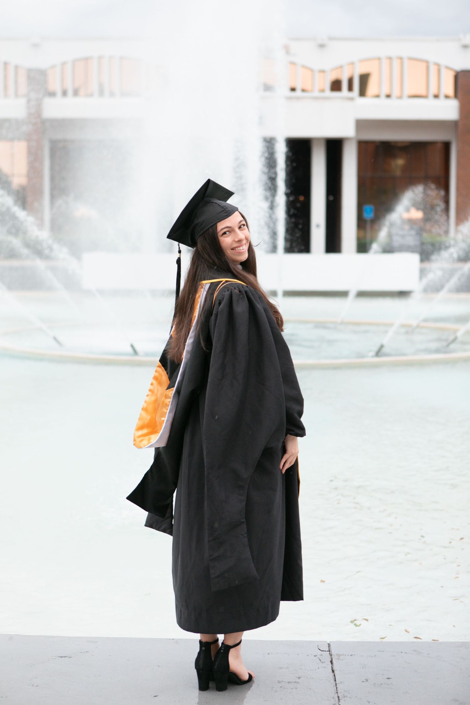 Smiling graduate in black cap and gown poses at UCF campus fountain.