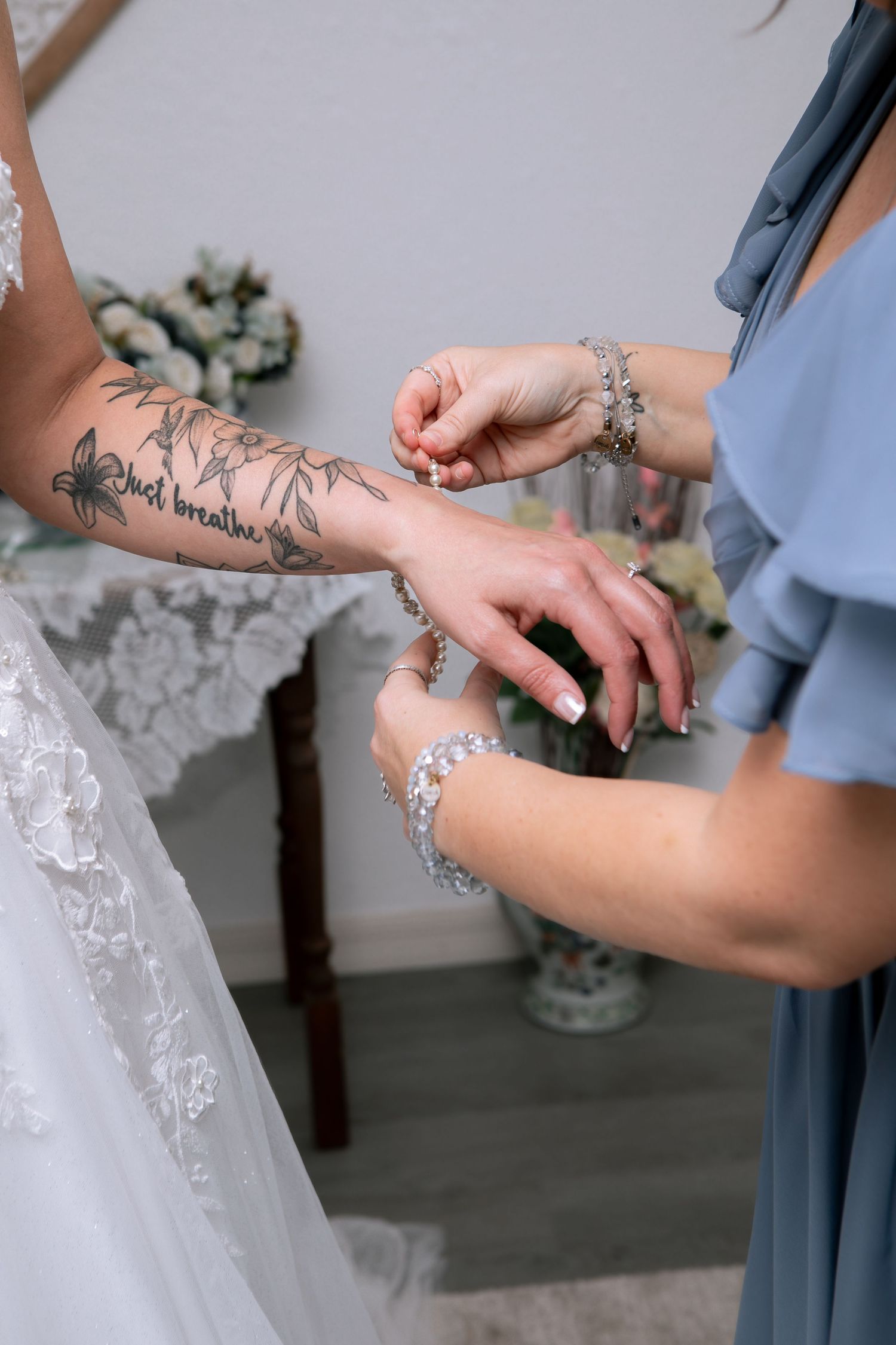 Bridesmaid helping bride put on a delicate crystal bracelet, bride has floral tattoo sleeve, both wearing elegant wedding attire.