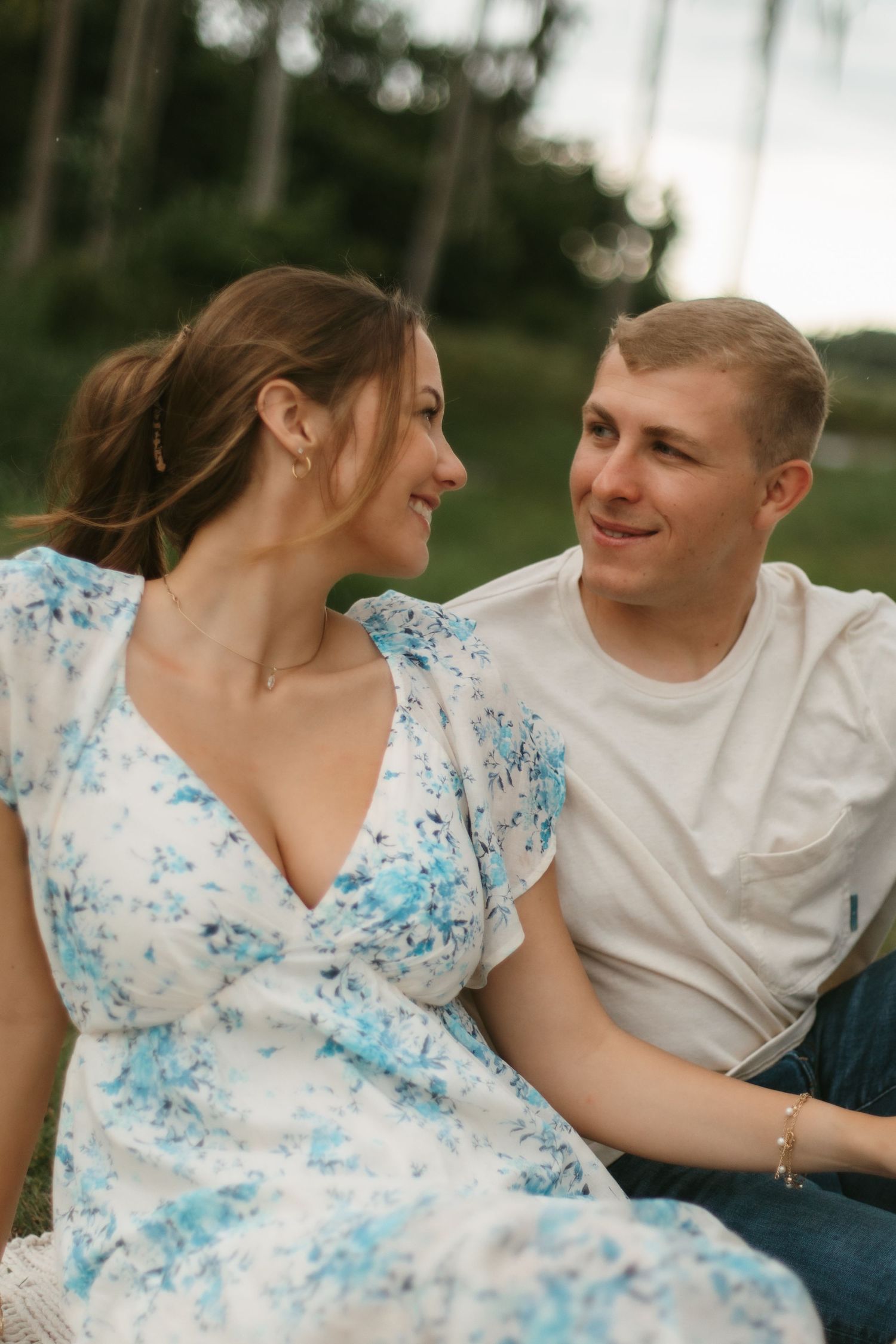 Smiling couple sitting outdoors gazing at each other, woman in white and blue floral dress, man in white t-shirt and jeans.