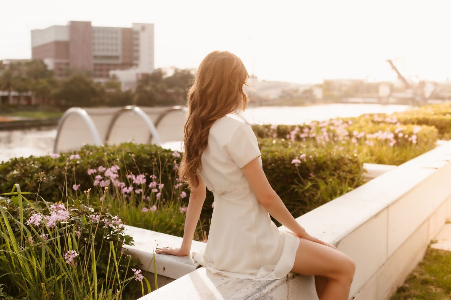 Young woman in white dress sitting on a garden wall at sunset, with flowers and a river in the background.