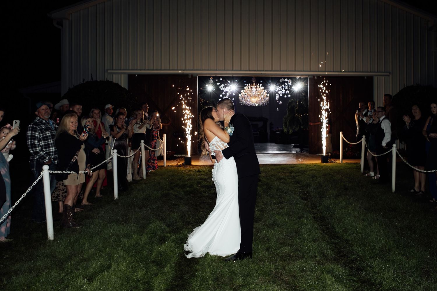 Bride and groom share a romantic kiss during sparkler exit at night outside a barn wedding venue.