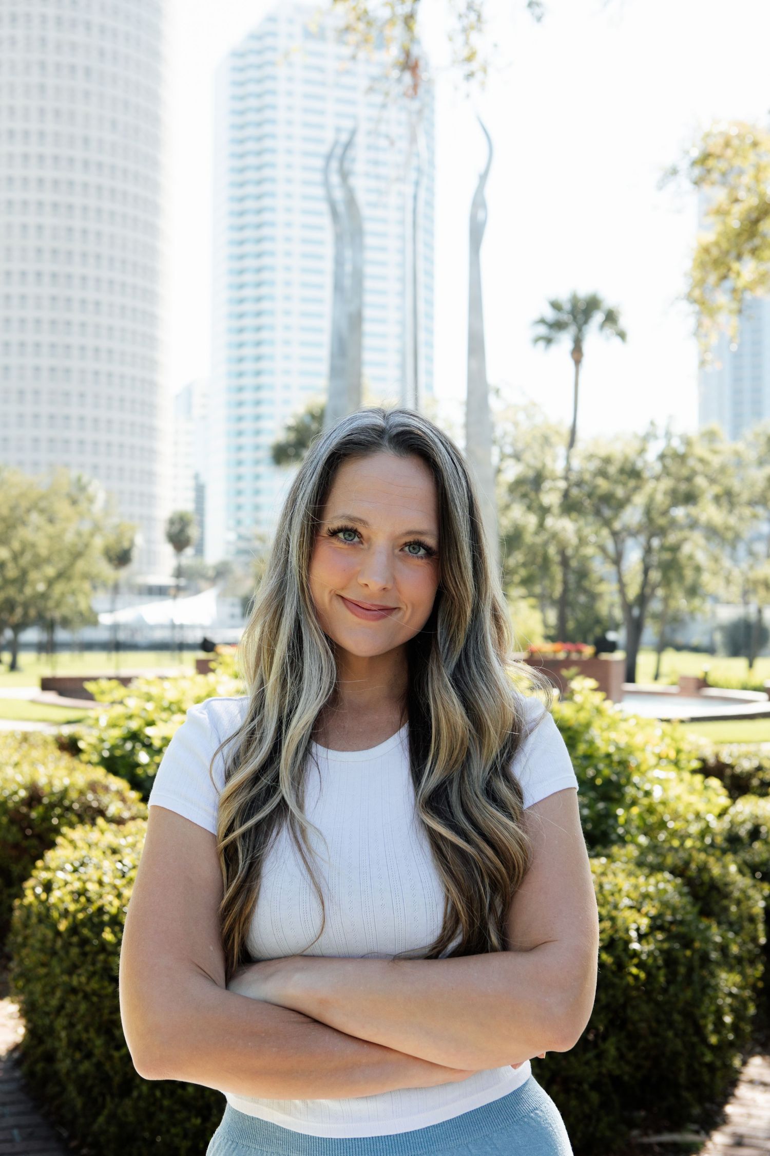 Confident young woman with long blonde hair standing arms crossed in urban park with city skyline.