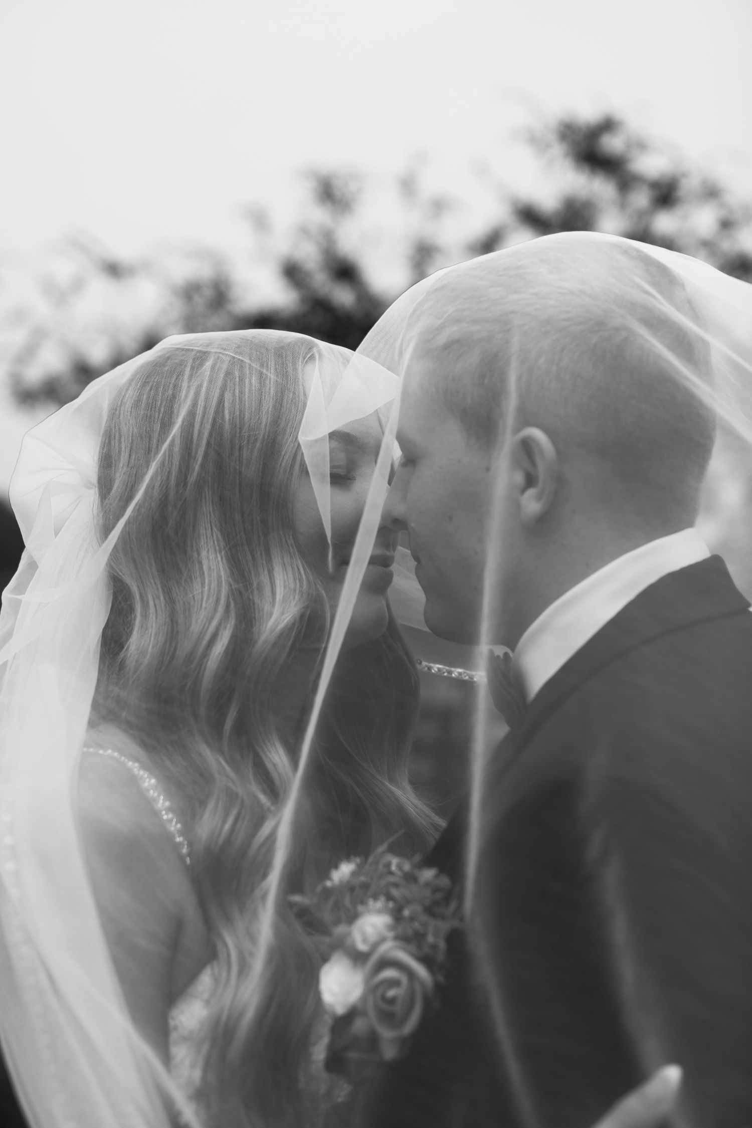Black and white photo of bride and groom kissing under wedding veil outdoors.
