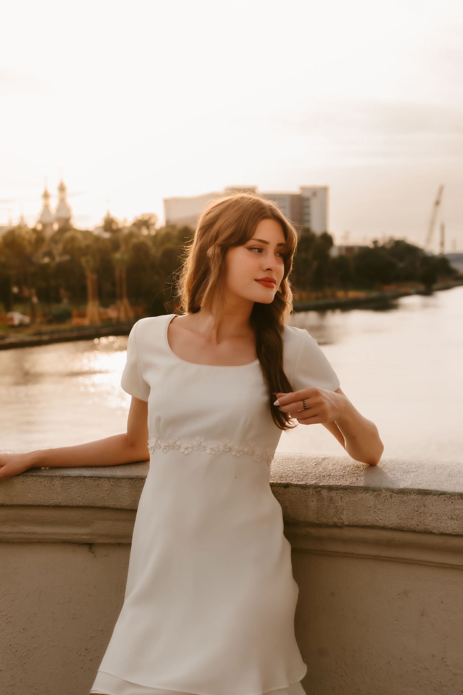 Young woman in white mini dress posing by a stone railing with a river and city skyline at golden hour.