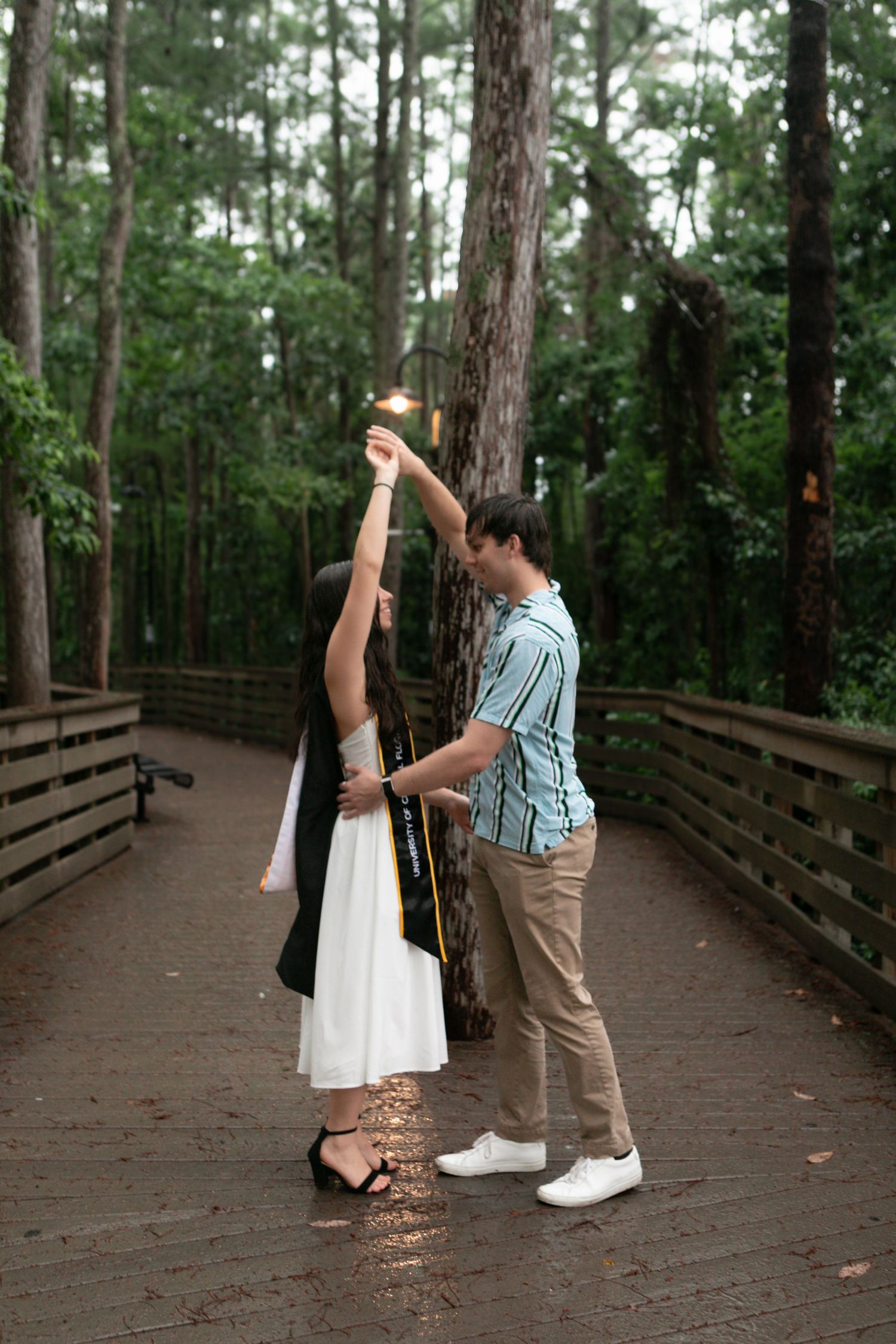 Couple dancing on a rainy wooden boardwalk surrounded by tall pine trees in a forest.