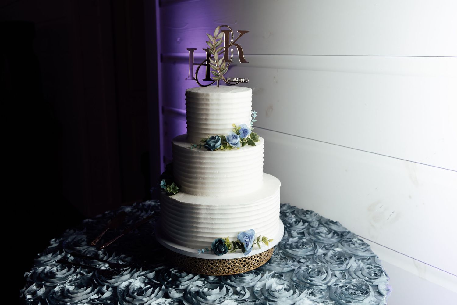 Three-tier white wedding cake with monogram topper and blue floral accents on decorative tablecloth.