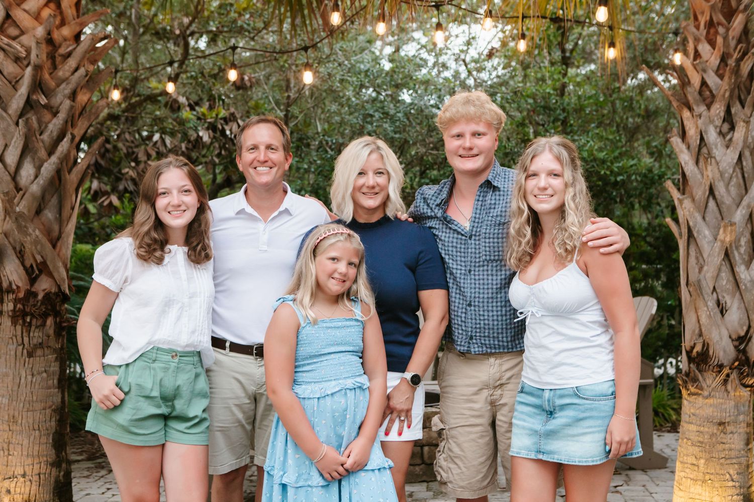 Family of six smiling together outdoors under string lights surrounded by tropical palm trees.