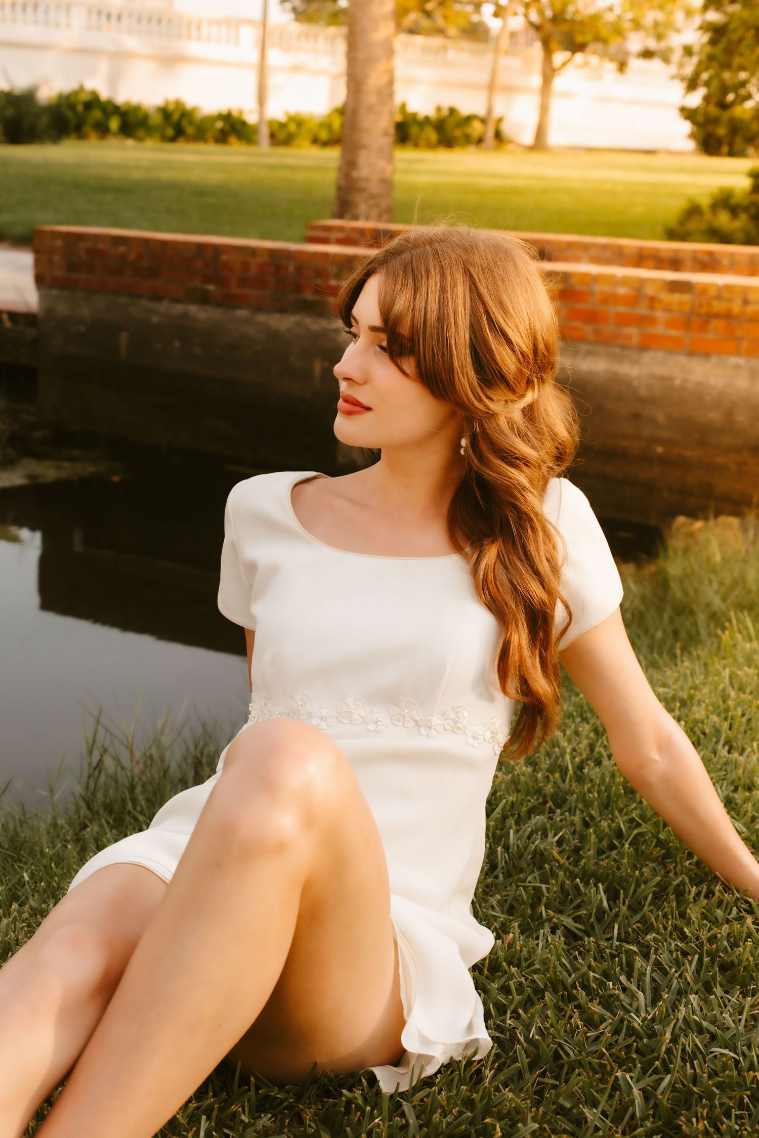 Woman in white mini dress sits on grass by pond at sunset, hair styled in soft waves, golden light glowing.