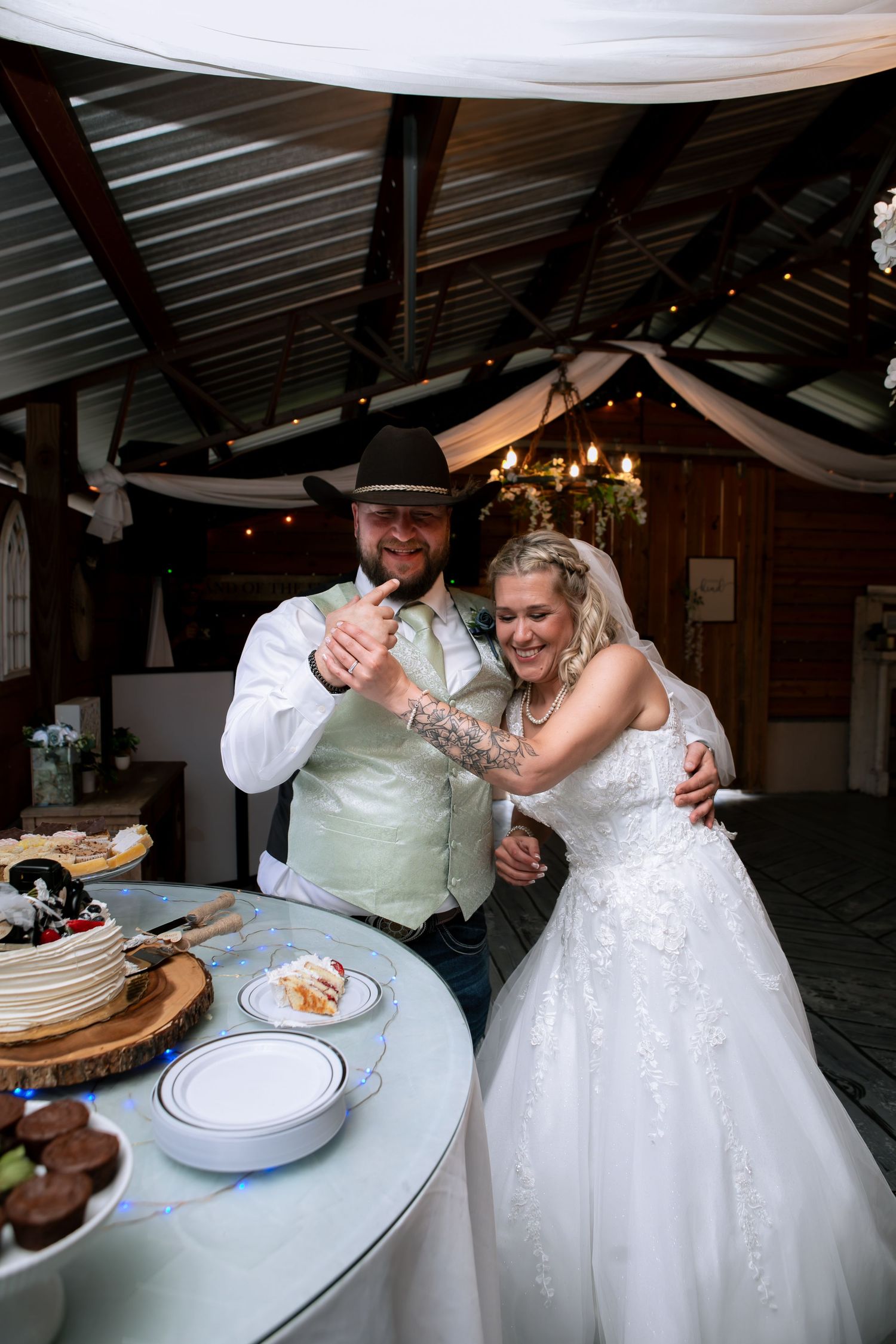 Smiling bride and groom in cowboy hat cut their wedding cake at a rustic barn reception venue.