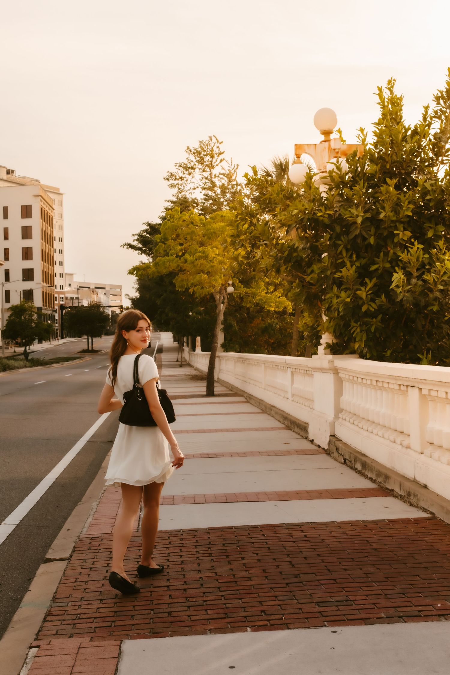 Young woman in white dress with backpack walking on a brick bridge sidewalk at golden hour.