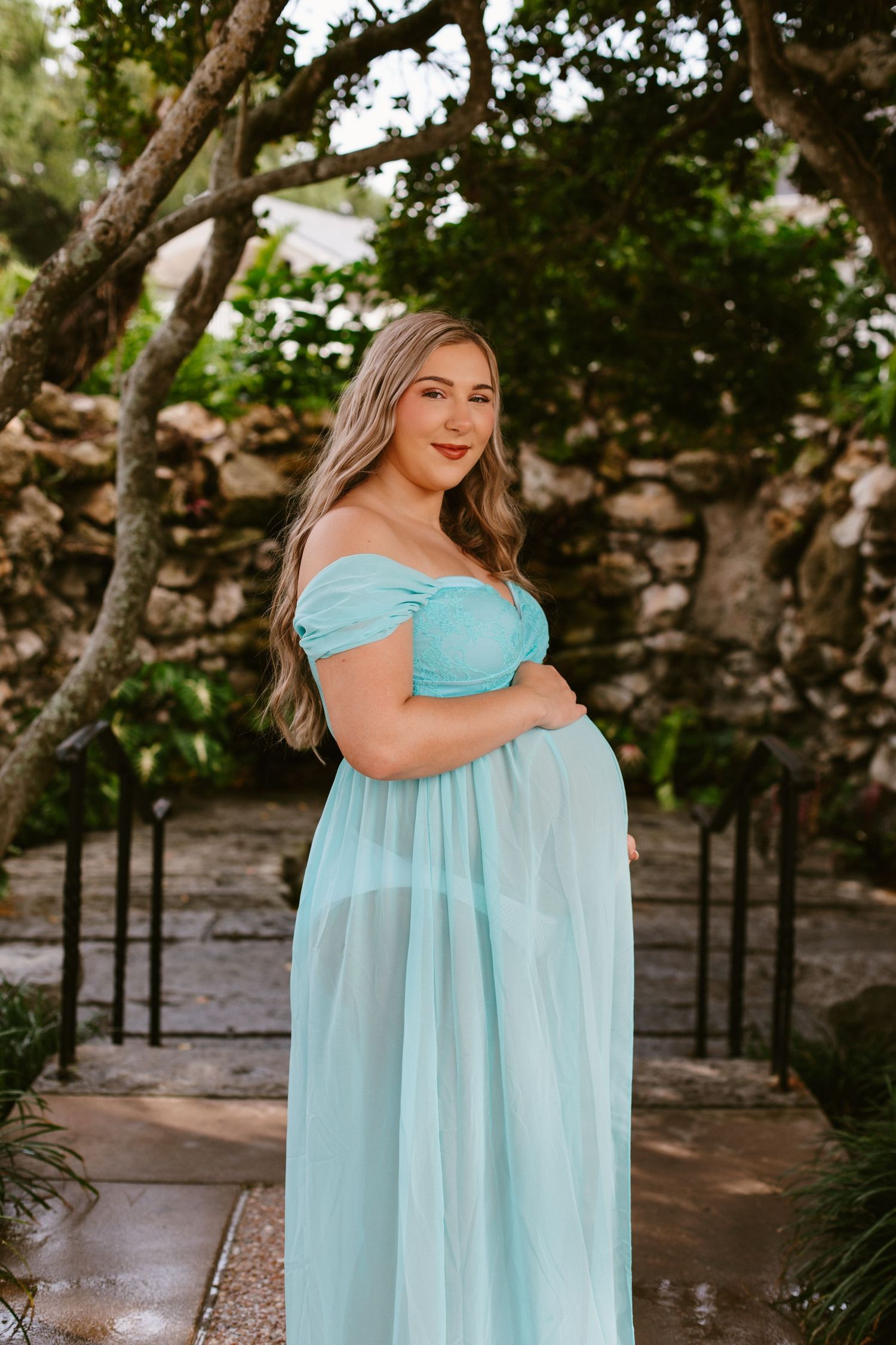 Pregnant woman in a light blue off-shoulder tulle maternity gown posing outdoors in a lush garden setting.