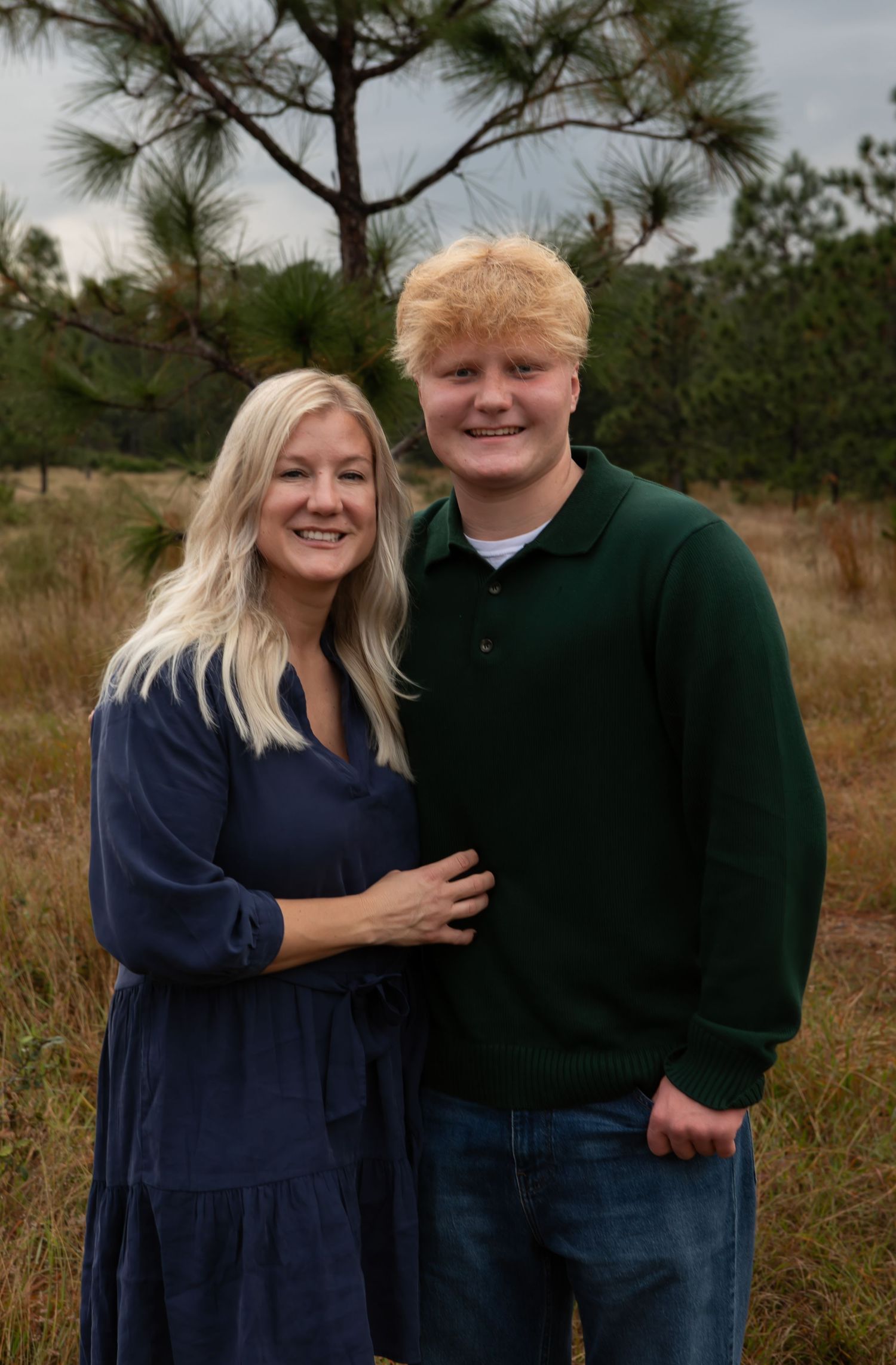 A smiling woman in a blue dress and a young man in a green sweater pose together outdoors among pine trees.