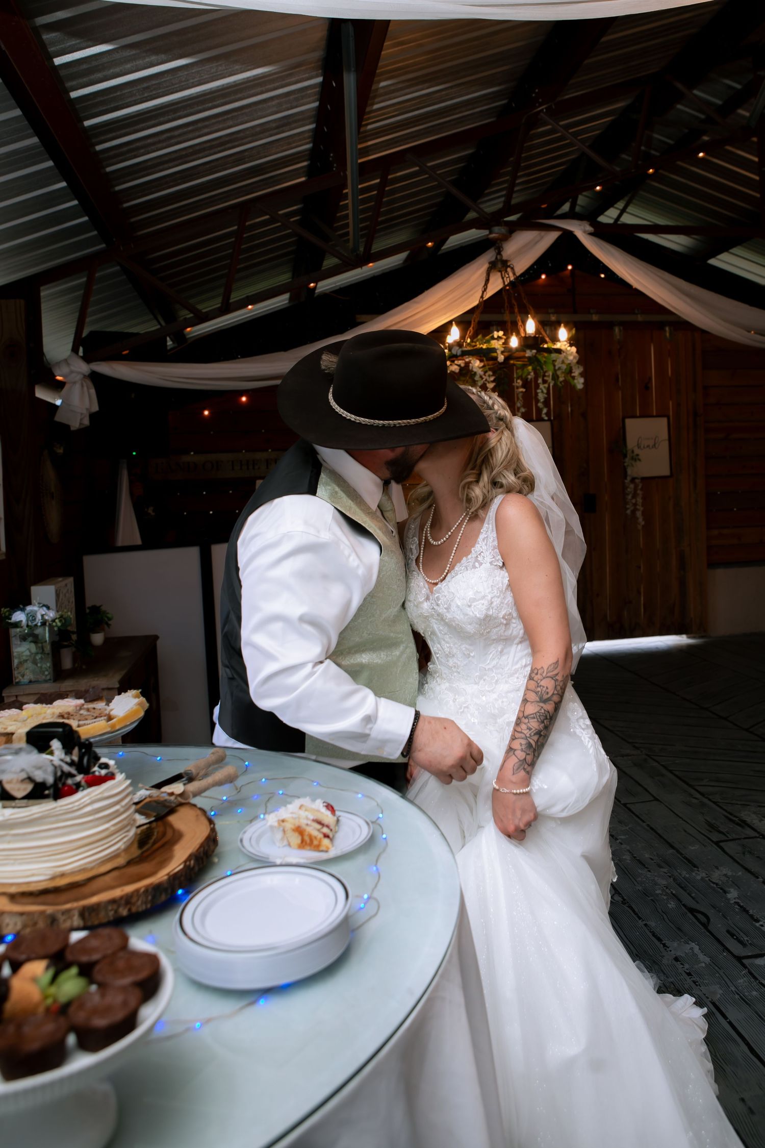 Groom in cowboy hat kisses bride in lace gown while cutting wedding cake at a rustic reception venue.