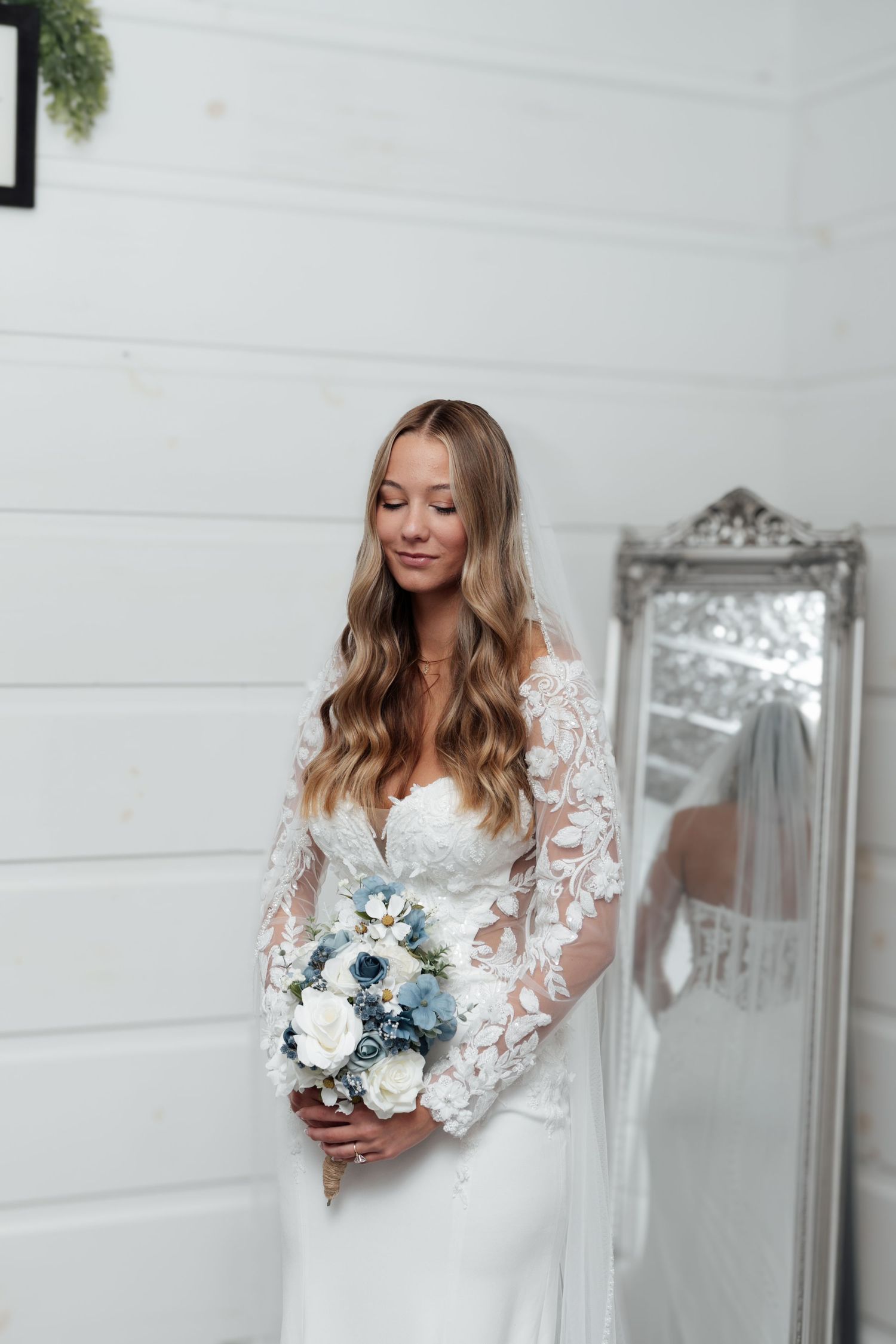 Bride in lace long-sleeve wedding gown holding blue and white bouquet, standing before ornate mirror in white room.
